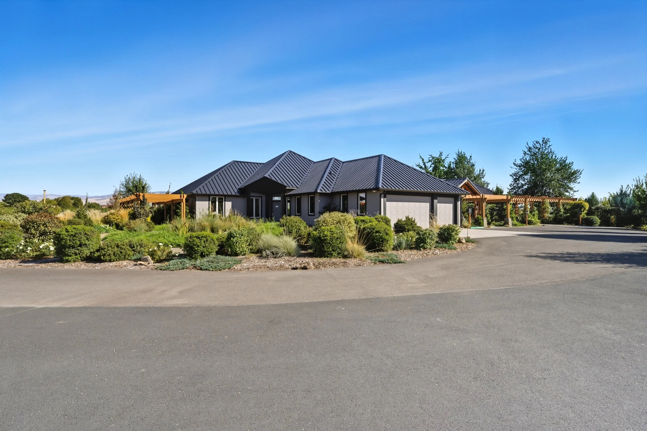 A modern single-story house with a dark metal roof, light gray walls, and attached garage, surrounded by a landscaped garden with bushes and plants, under a clear blue sky.