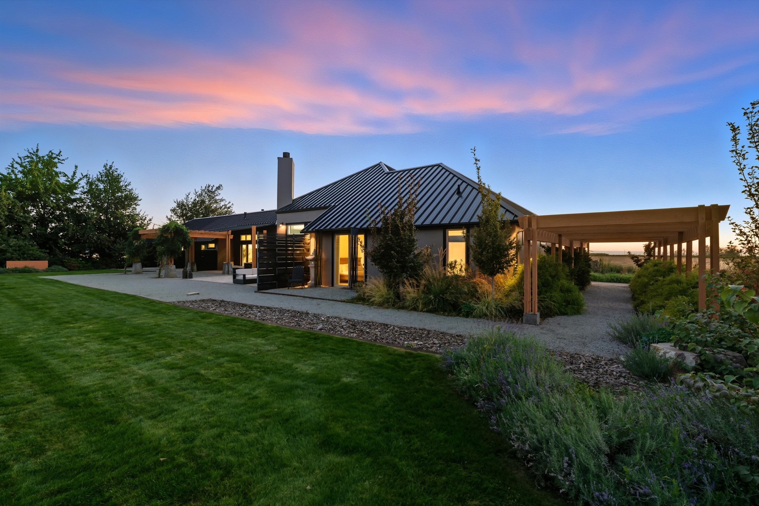 Modern house with black metal roof and large windows, surrounded by landscaped garden and patio furniture at sunset.