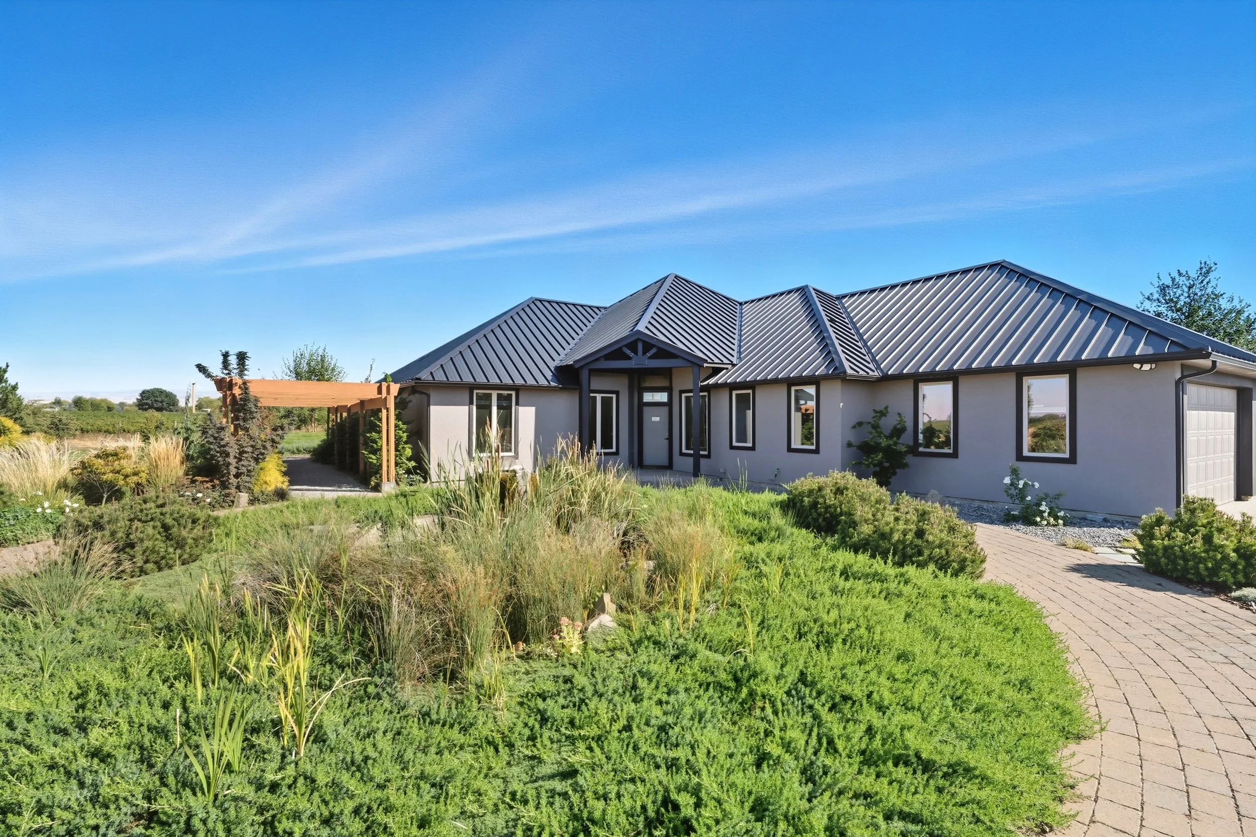 A modern single-story house with a metal roof, surrounded by a well-maintained garden and a curved brick pathway in clear weather.
