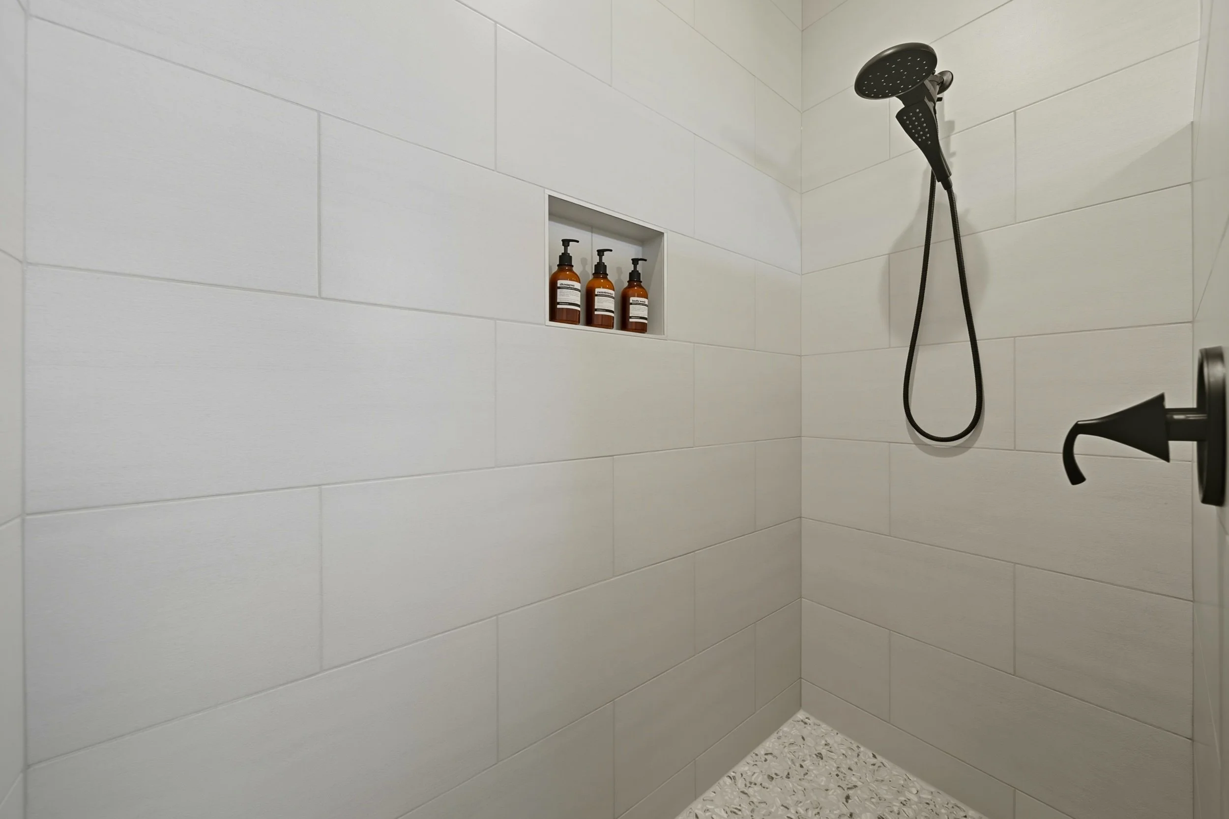 A minimalist walk-in shower with beige tiled walls, a black handheld showerhead, a built-in niche with three amber soap bottles, and a black wall-mounted faucet handle.