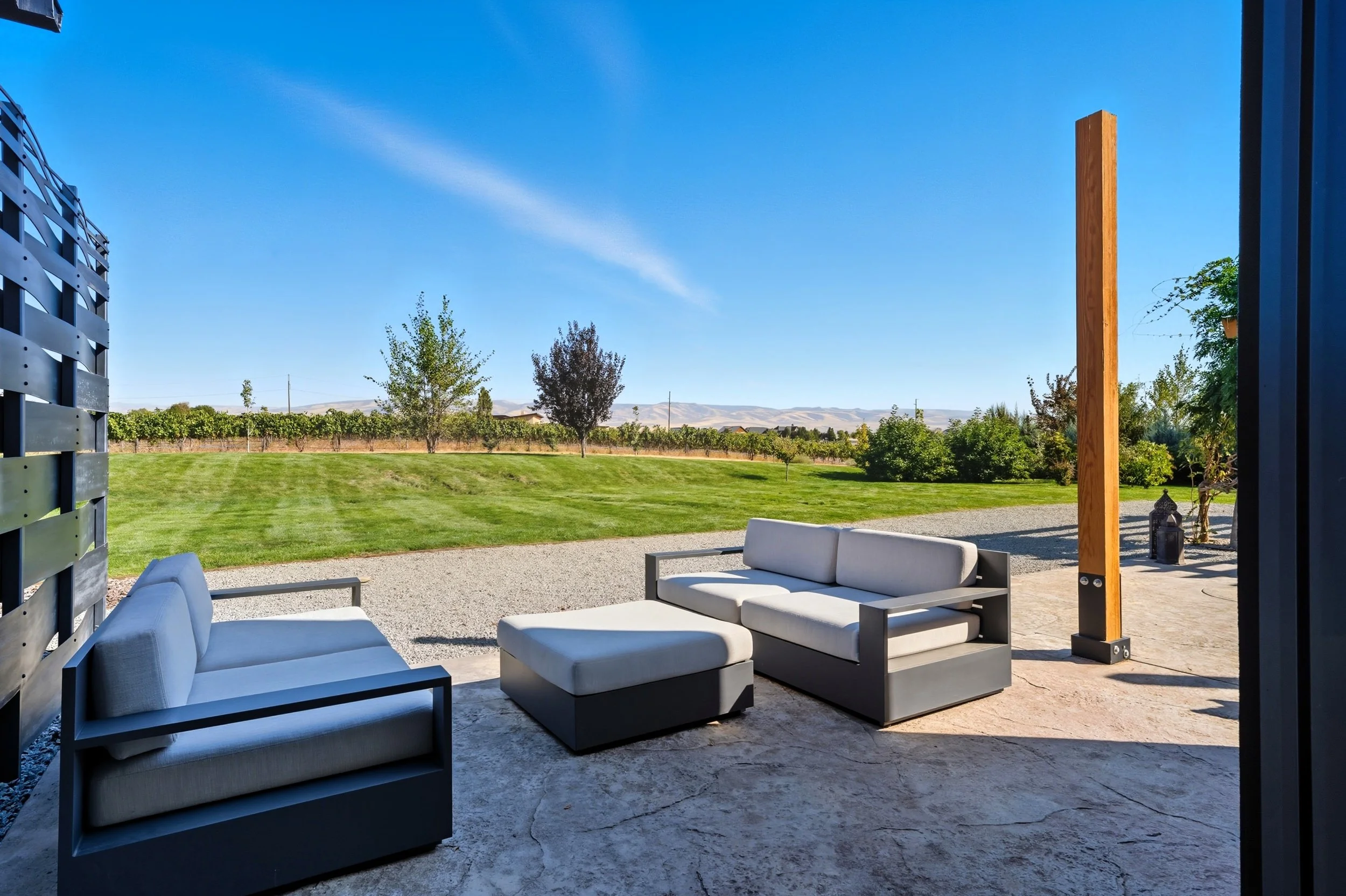 Outdoor patio with modern white cushioned seating, a grey ottoman, and a wooden post with a golf driving net, overlooking a green lawn with trees and mountains in the background under a clear blue sky.