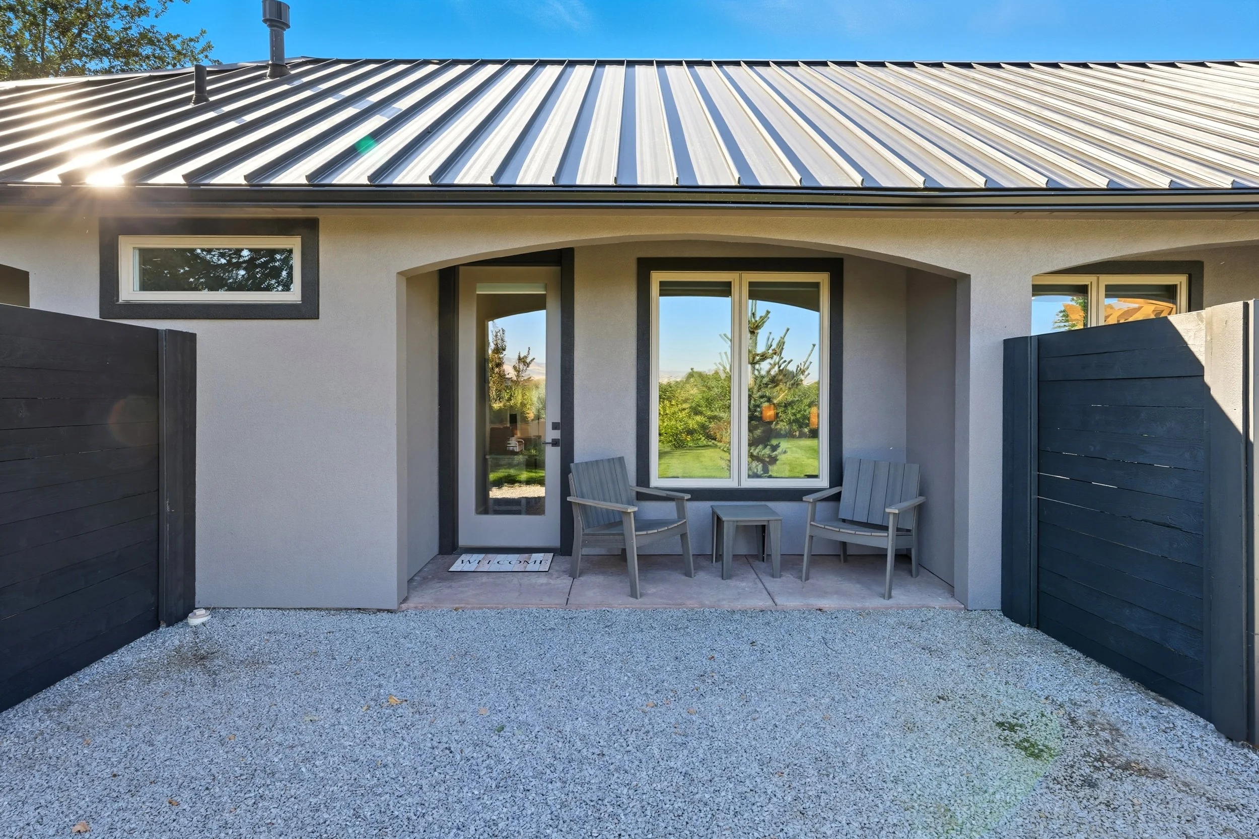 Front porch of a house with a small seating area and a window, surrounded by a gravel yard and a black privacy fence.