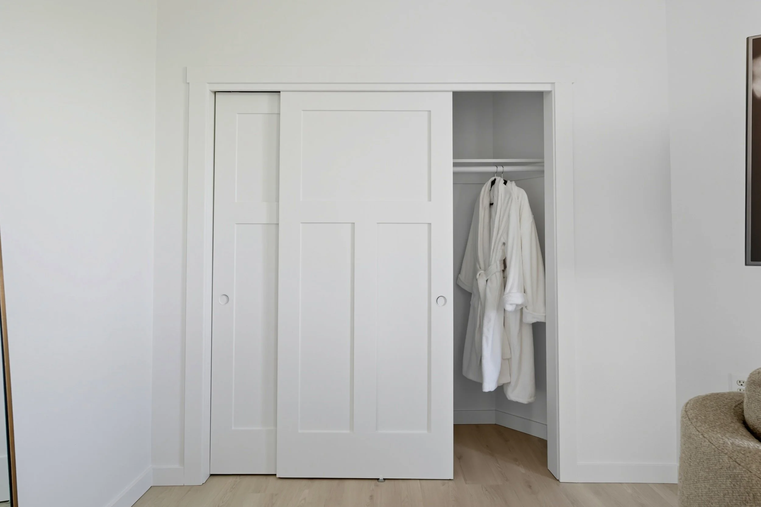 white closet with sliding doors, one door open revealing a bathrobe hanging inside, with a light-colored wall and hardwood floor