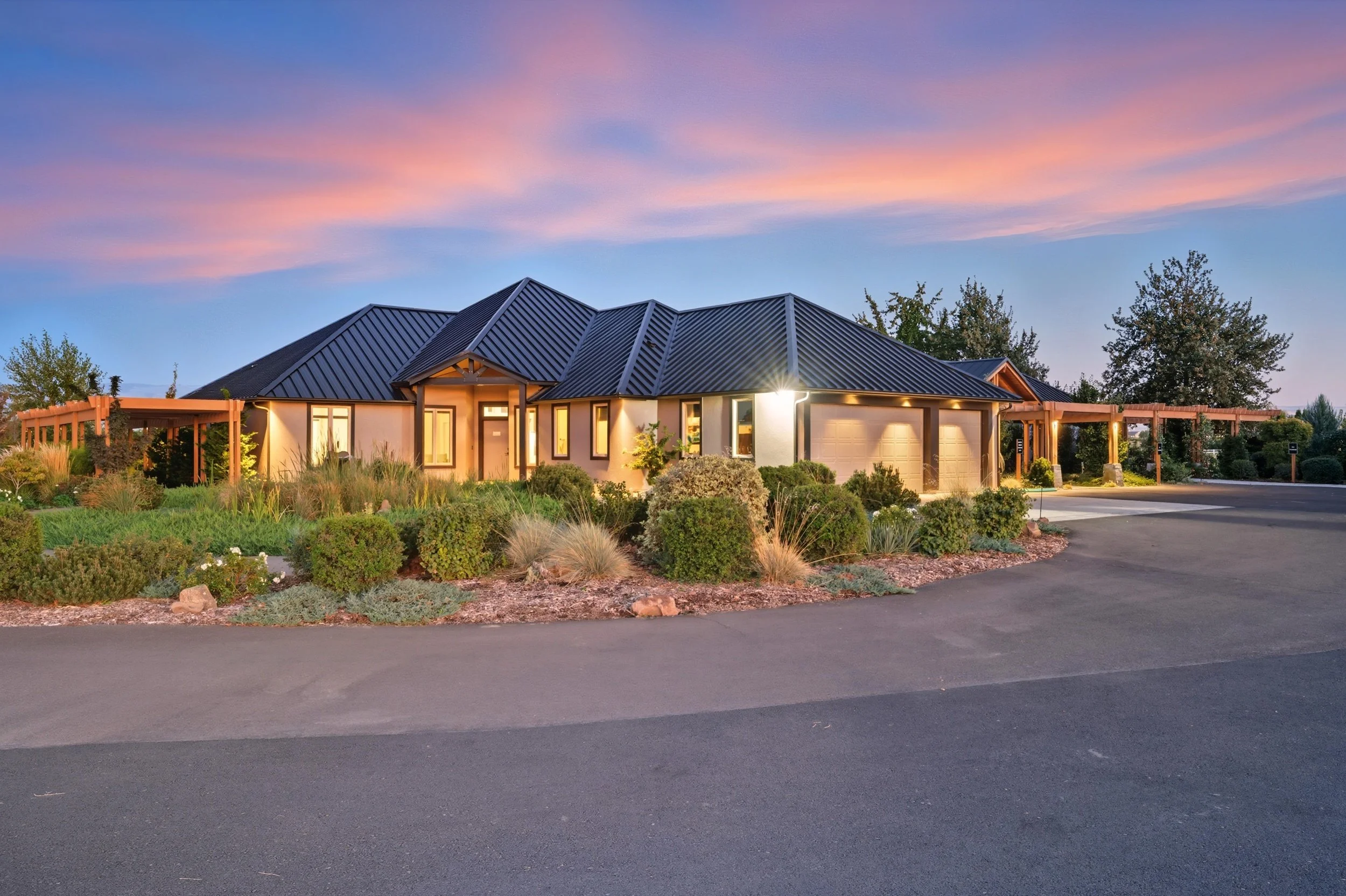 A modern house with a black metal roof, surrounded by landscaped garden and shrubbery at sunset.