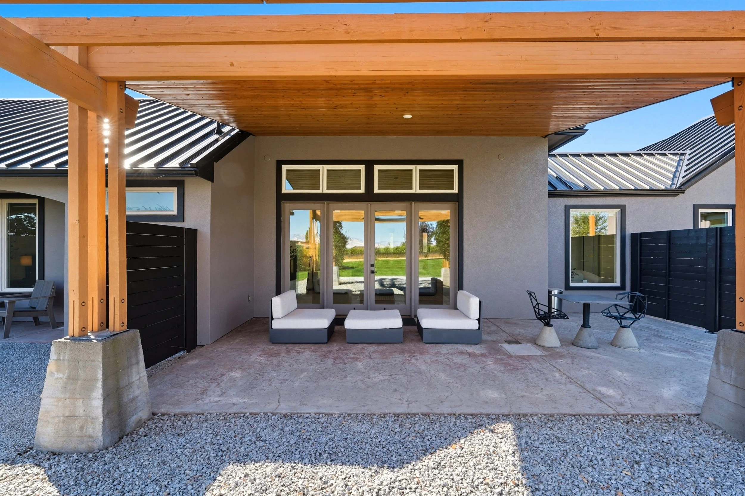 Modern outdoor patio with white cushioned furniture, concrete flooring, and a wooden pergola, overlooking a green lawn and a blue sky.