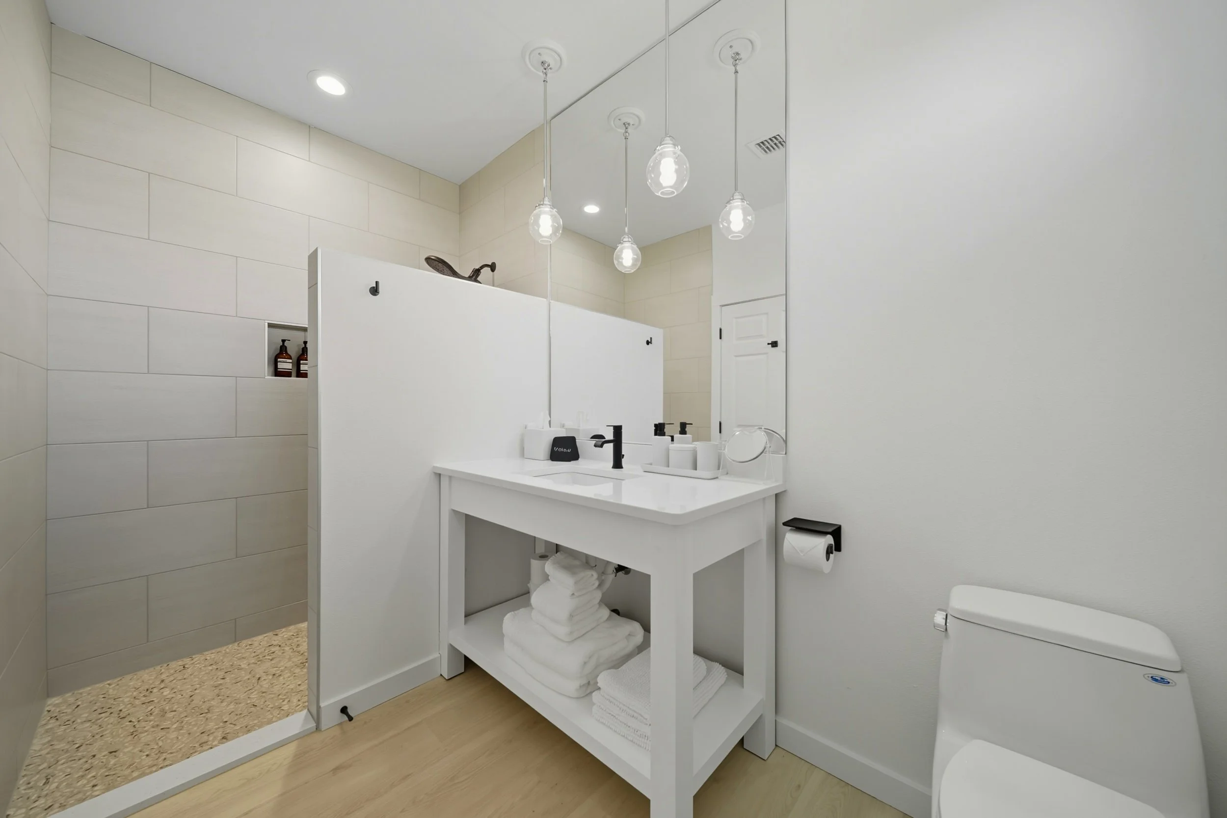 Modern bathroom with a white vanity, towels, mirror, toilet, shower area with a partial wall, and pendant lights.