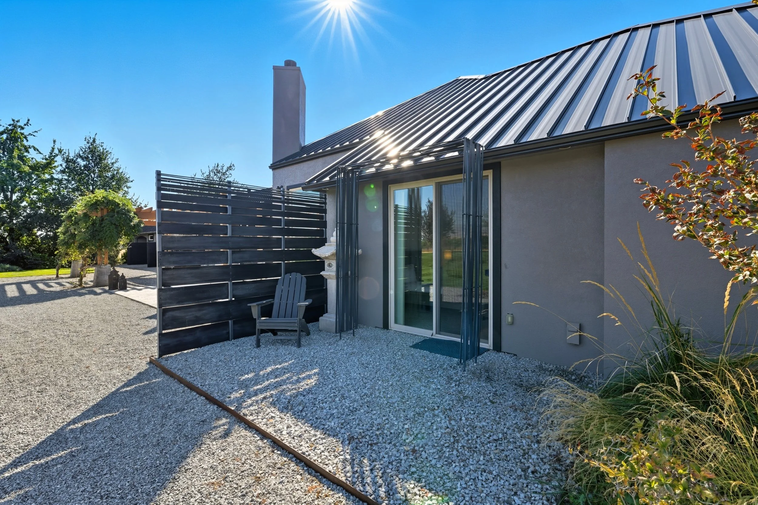 Backyard patio with gravel ground, gray Adirondack chair, black privacy fence, house with sliding glass door, metal roof, and a chimney, sun shining with lens flare