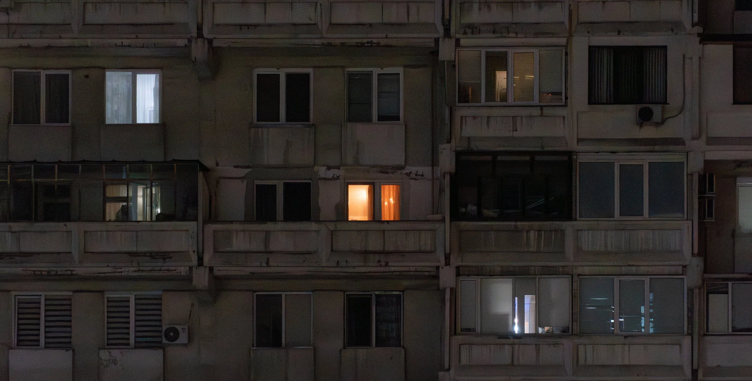Night view of a multi-story residential building with several windows, some lit, revealing interior rooms, balconies, and exterior air conditioning units.