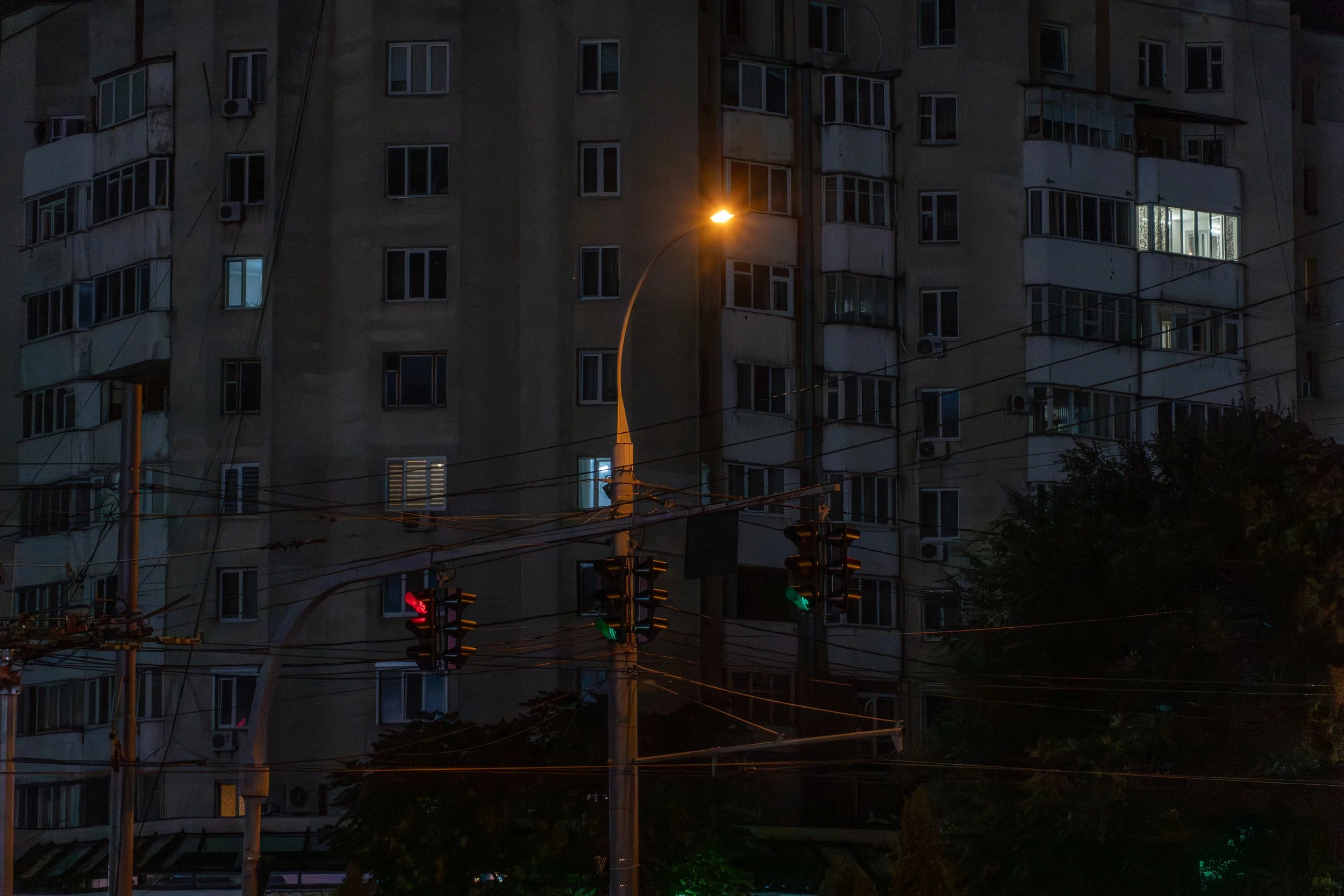 Nighttime city scene showing an apartment building with lit windows, streetlights, and traffic signals.
