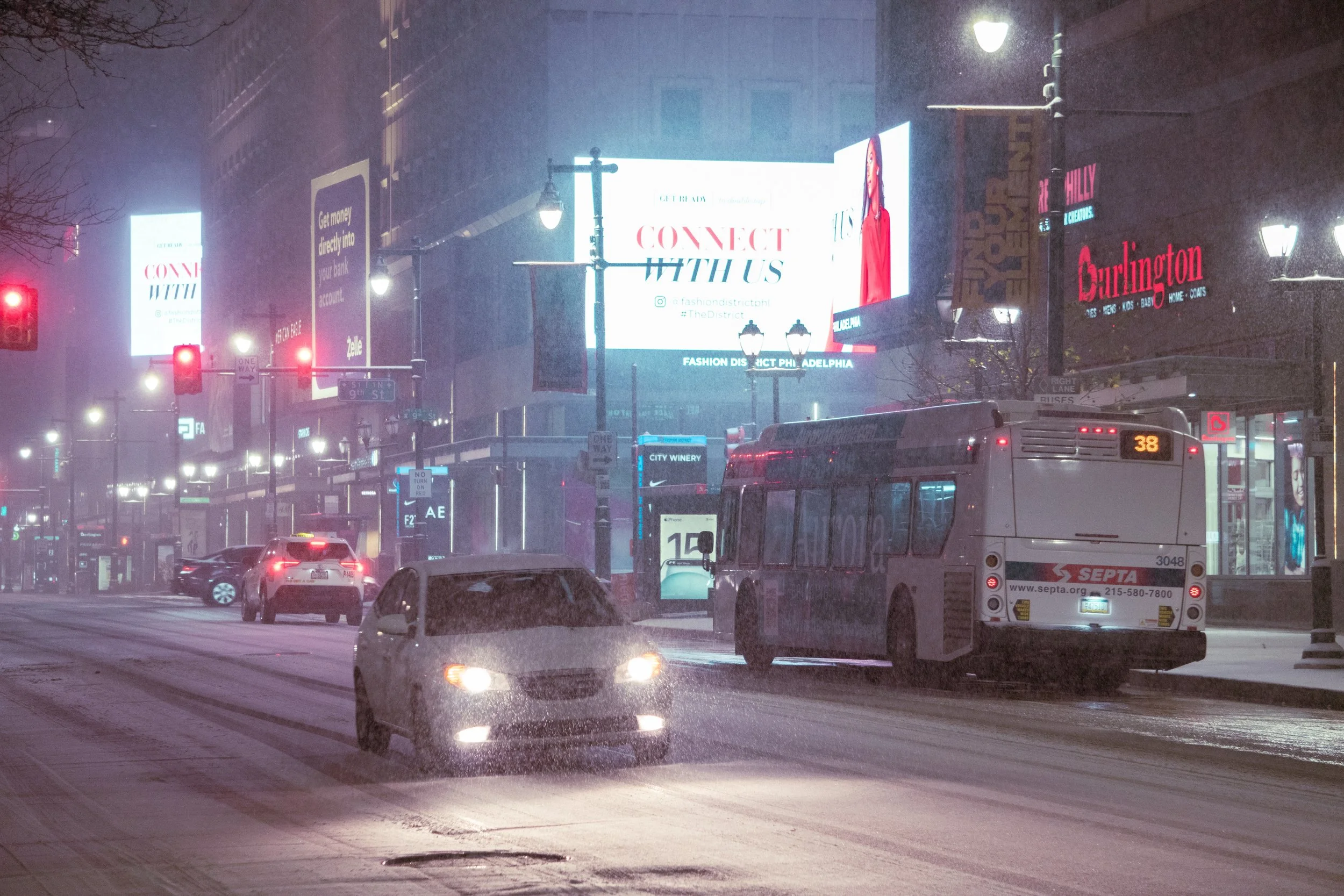 A city street at night with snow falling, showing cars and a bus, lit up billboards, and streetlights.