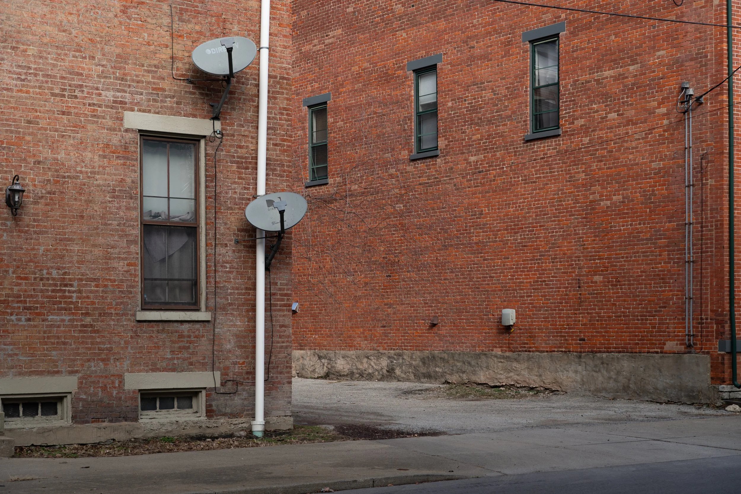 View of a brick building with three small windows and two satellite dishes attached to the outside wall, with a sidewalk and an alleyway in front.