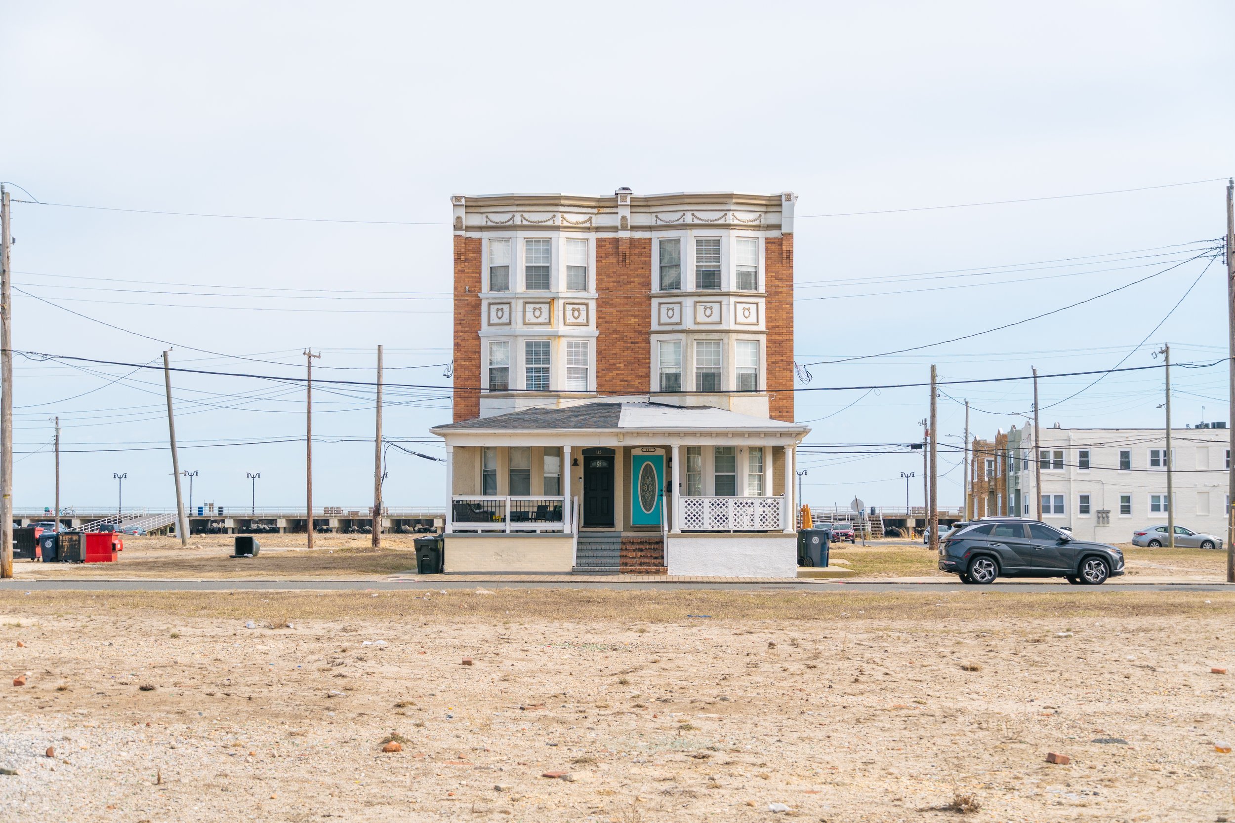 A multi-story house with Victorian architecture style, featuring a porch with white railings, a green front door, and multiple bay windows with intricate decorative details. There are power lines and cars in the background, and the house is situated on a somewhat barren, sandy lot.
