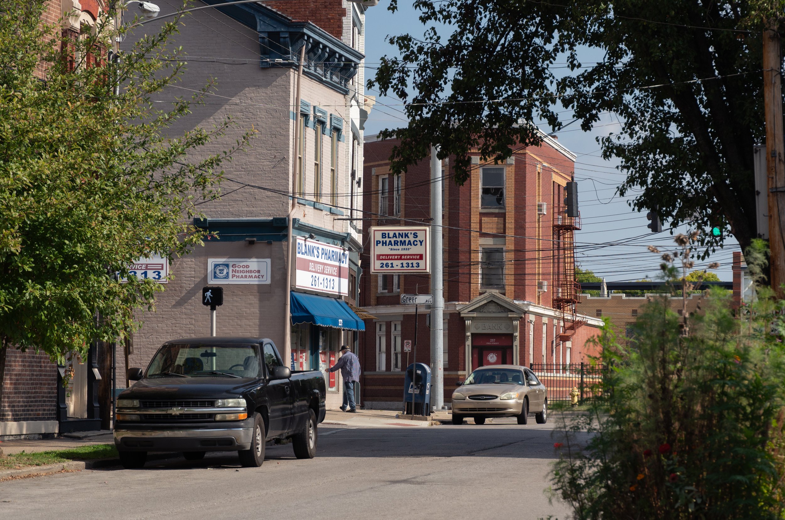 A street scene with a beige building housing Blank's Pharmacy and a man entering the store. There are parked cars, trees, and overhead power lines. The street sign indicates Greer Ave.