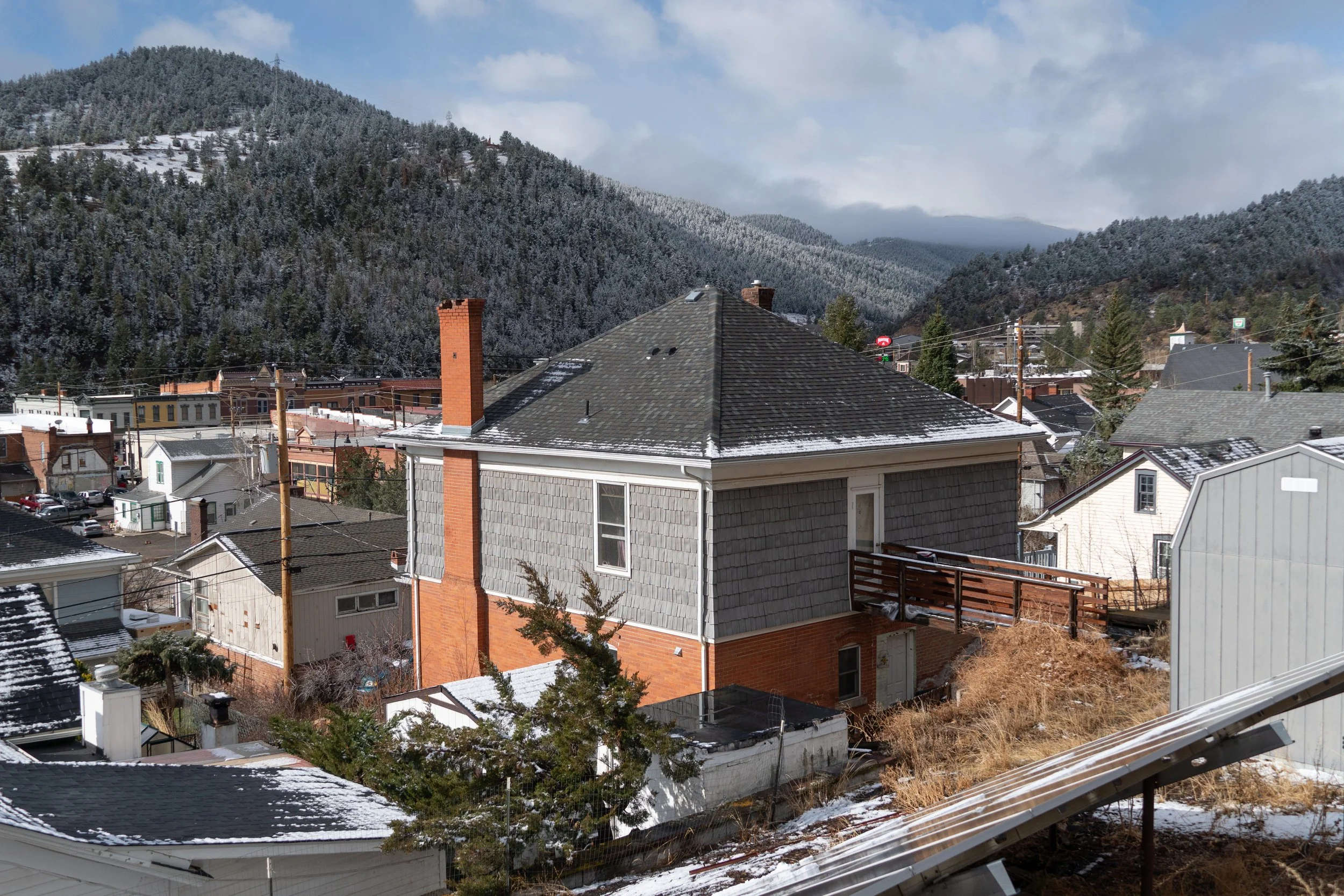 Residential neighborhood with houses, some snow on rooftops, mountains in the background, and cloudy sky.