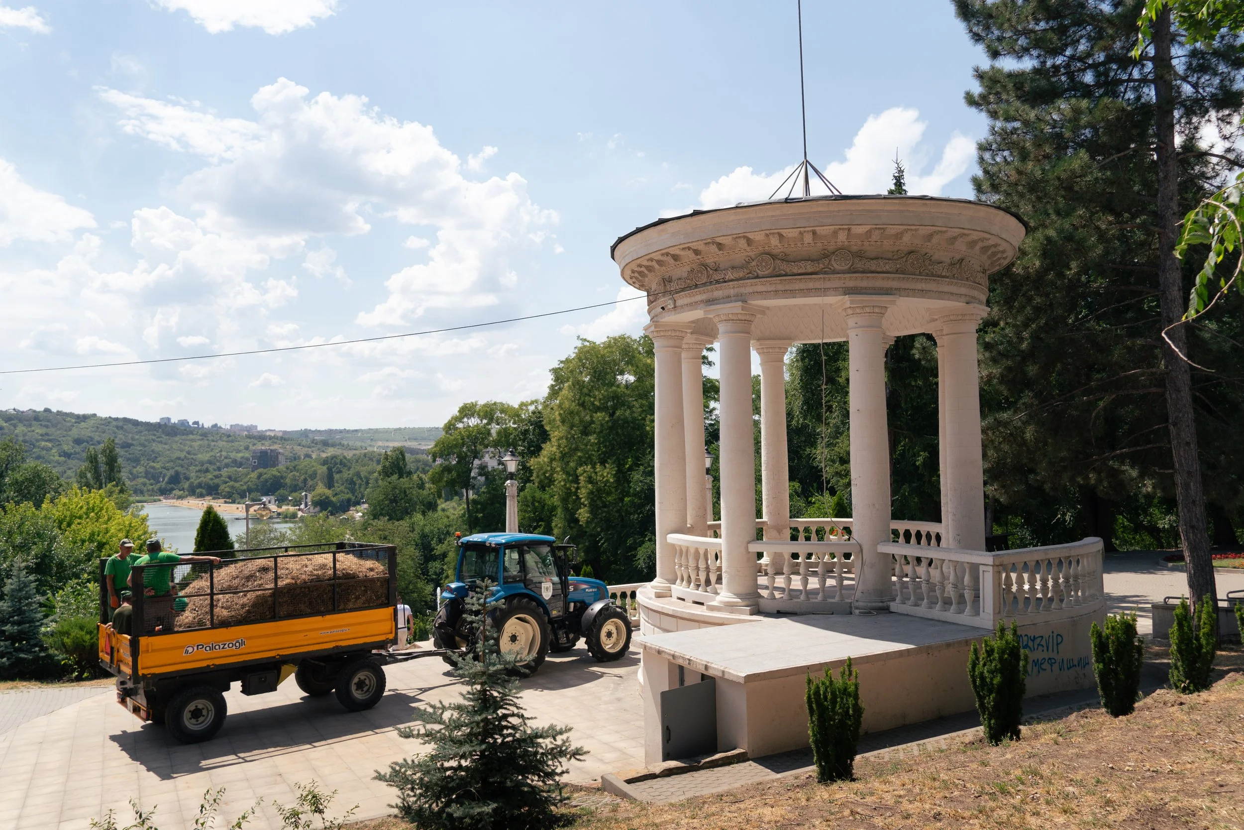 A scenic outdoor scene with a white classical-style pavilion with columns, a tractor pulling a trailer loaded with hay, and two people riding in the trailer near a river with lush greenery and blue sky with clouds.