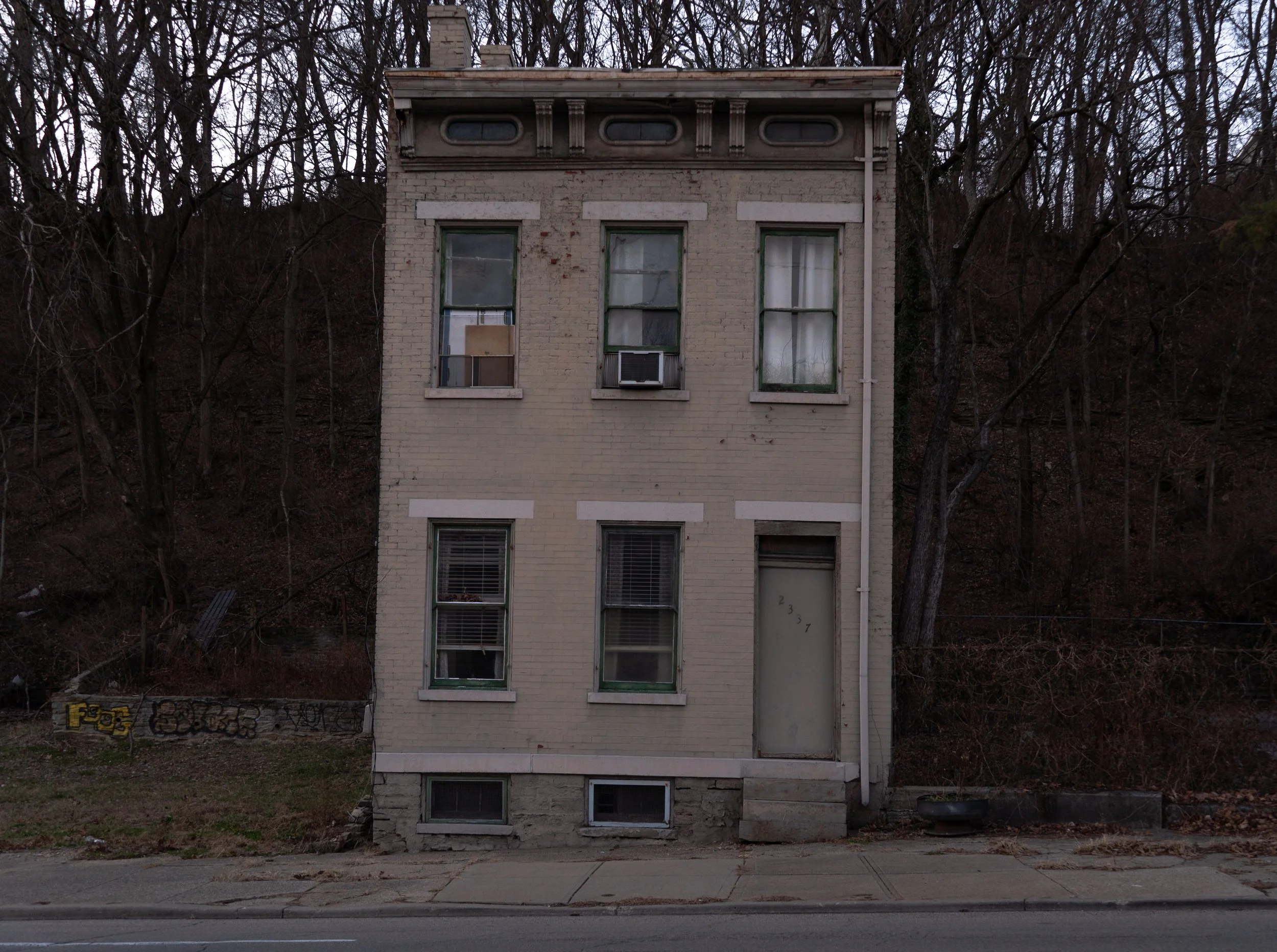 A three-story old brick building with six windows and a door, situated on a sidewalk with a hillside covered in leafless trees in the background.