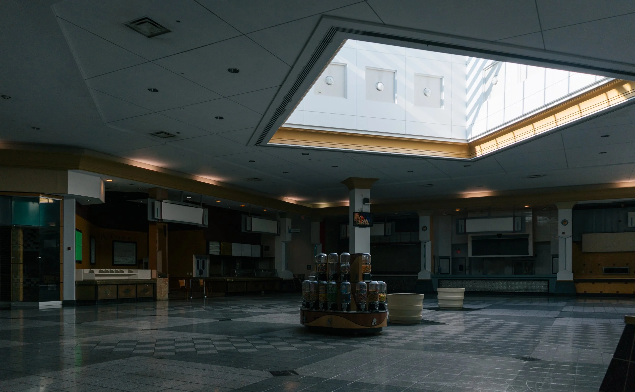 Empty indoor shopping mall or food court area with closed shops, a central display of gumball machines, and a large skylight ceiling.