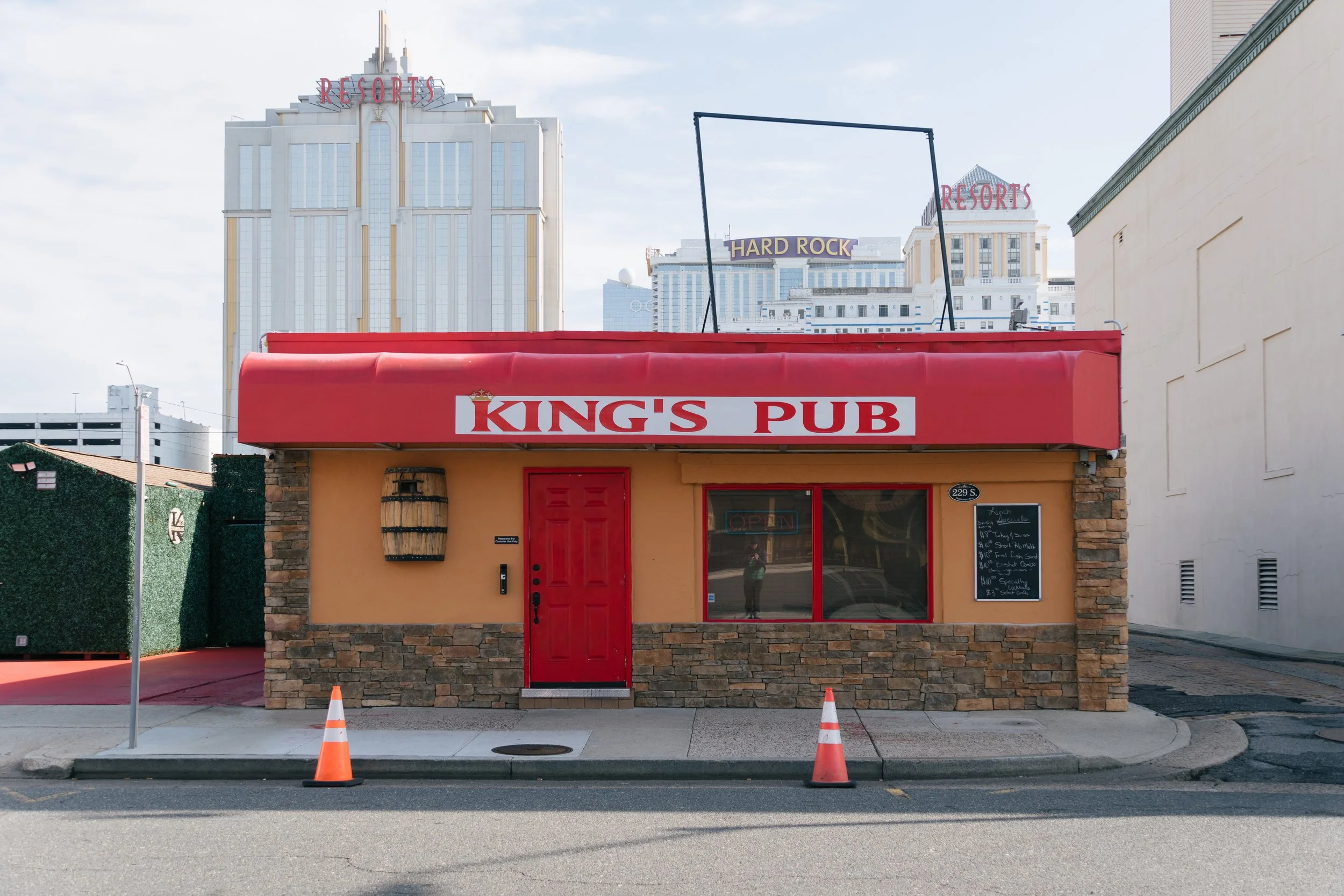 Small pub building with a red awning labeled 'King's Pub', a red door, a window with an 'Open' sign, and a chalkboard menu outside. Traffic cones are in front of the curb. In the background, tall buildings and signs for Resorts and Hard Rock are visible.