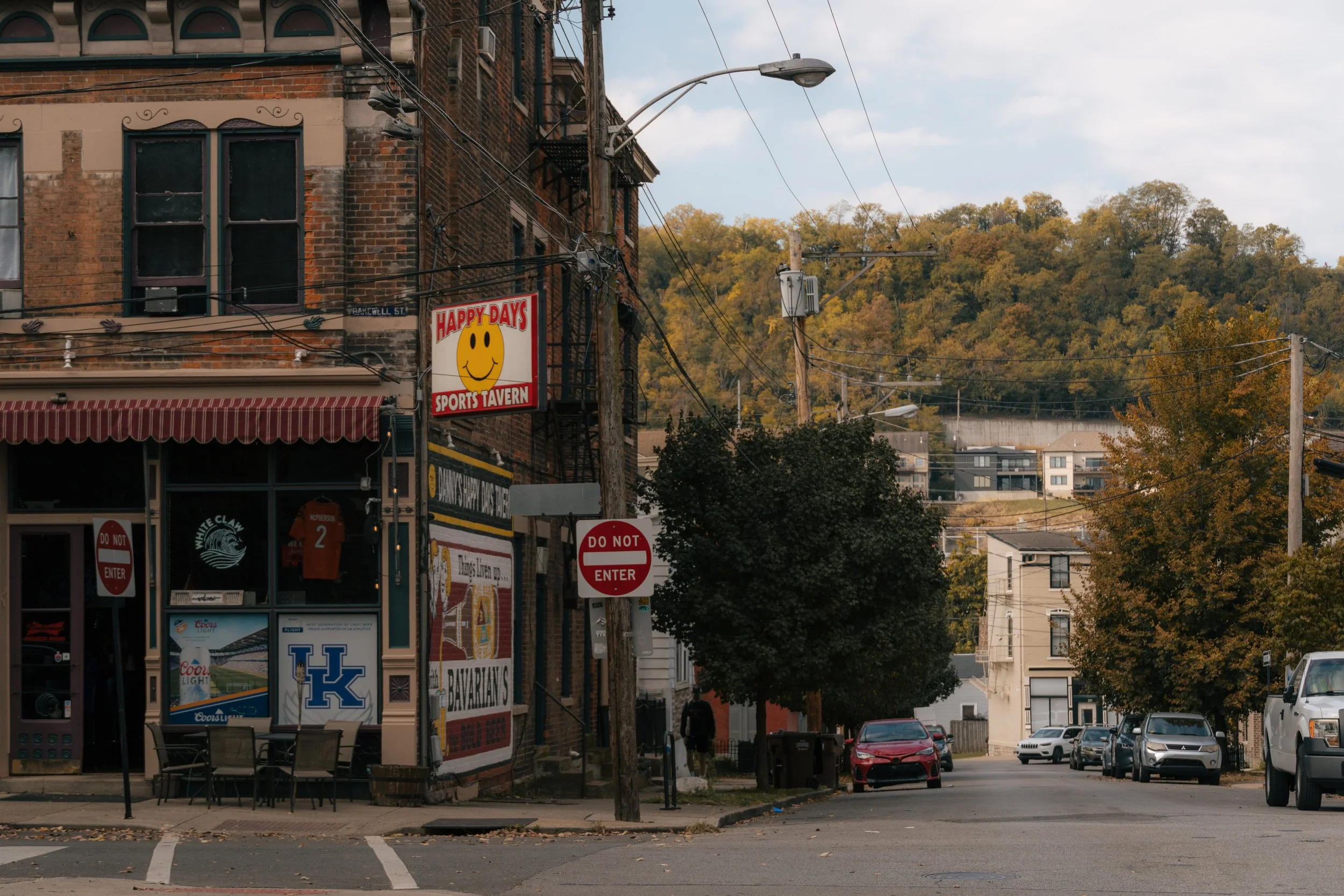 Street view with a sports tavern named Happy Days featuring a sign with a yellow smiley face. The area has parked cars, trees, and a hilly background with buildings and utility lines.