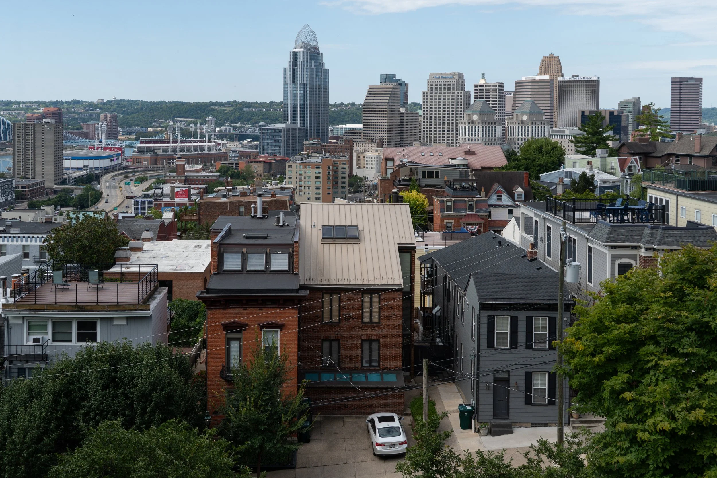 View of a city skyline with tall buildings, houses with rooftops, and trees in the foreground, under a partly cloudy sky.
