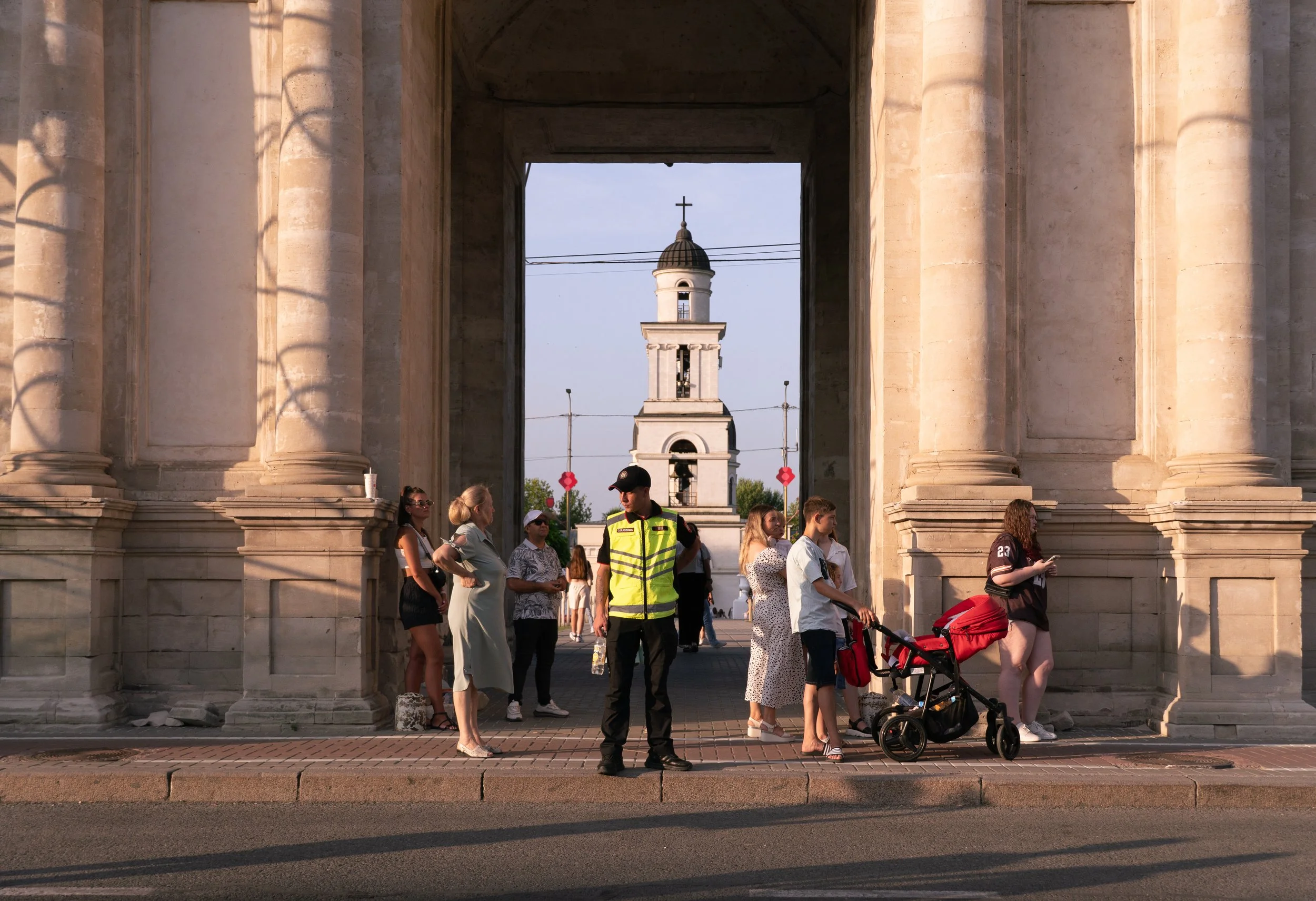 People gathered under a large stone archway with a view of a bell tower in the background, and a police officer standing in front.