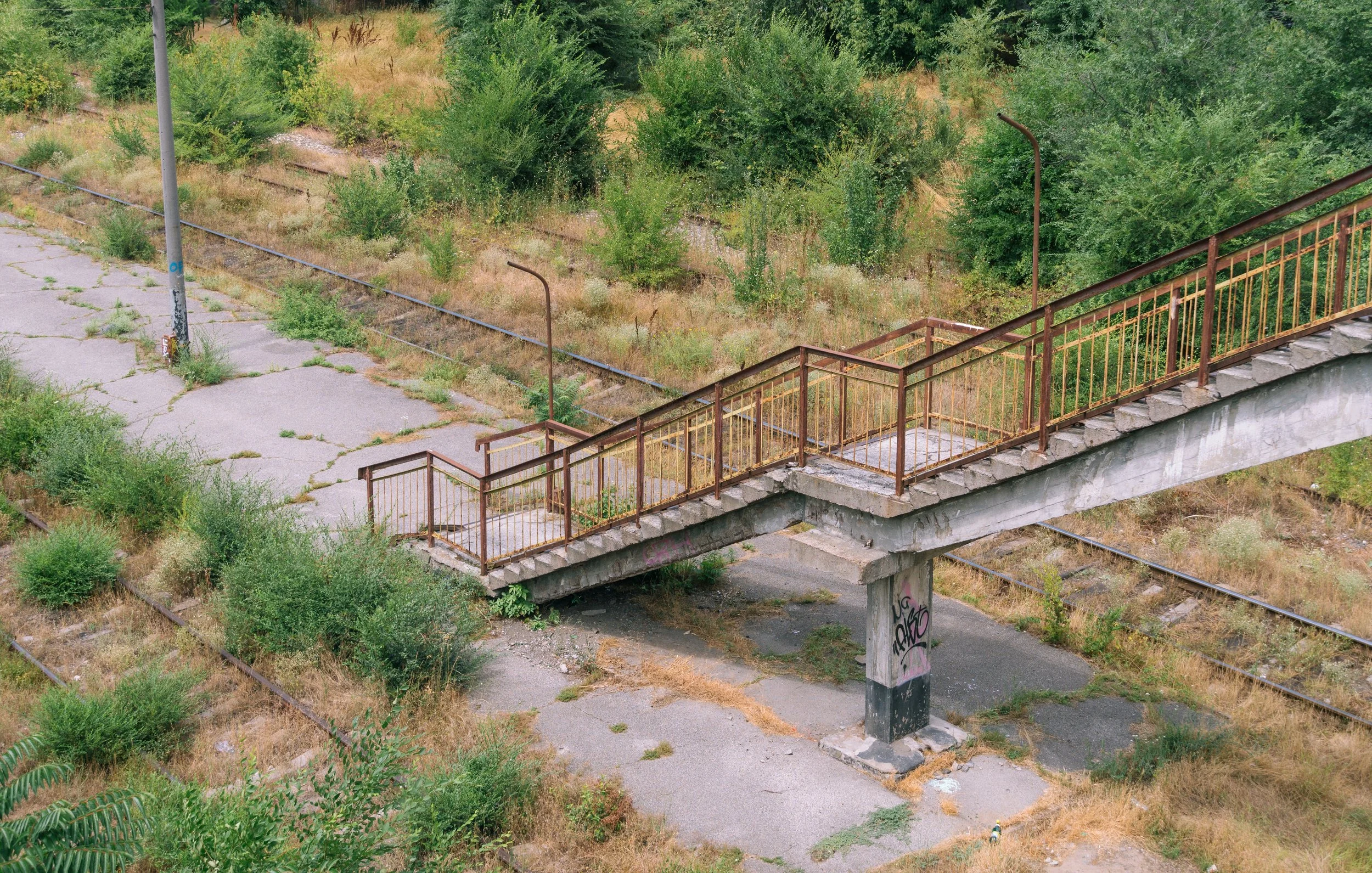 An abandoned pedestrian bridge with rusty railings over a concrete support, surrounded by overgrown grass, weeds, and cracked asphalt, with train tracks running parallel underneath, and a wooded area in the background.