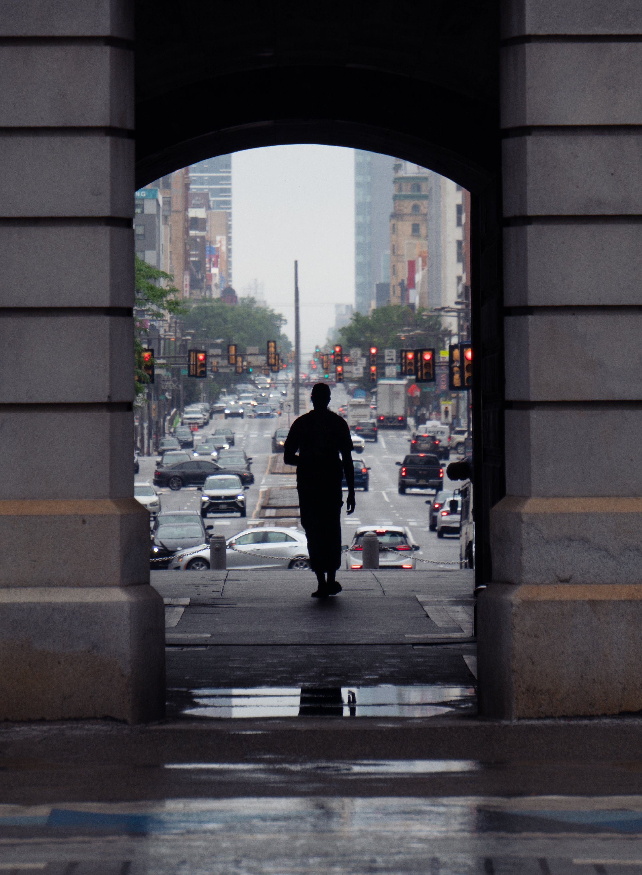 Silhouette of a person walking through an archway onto a busy city street with traffic lights and buildings in the background.