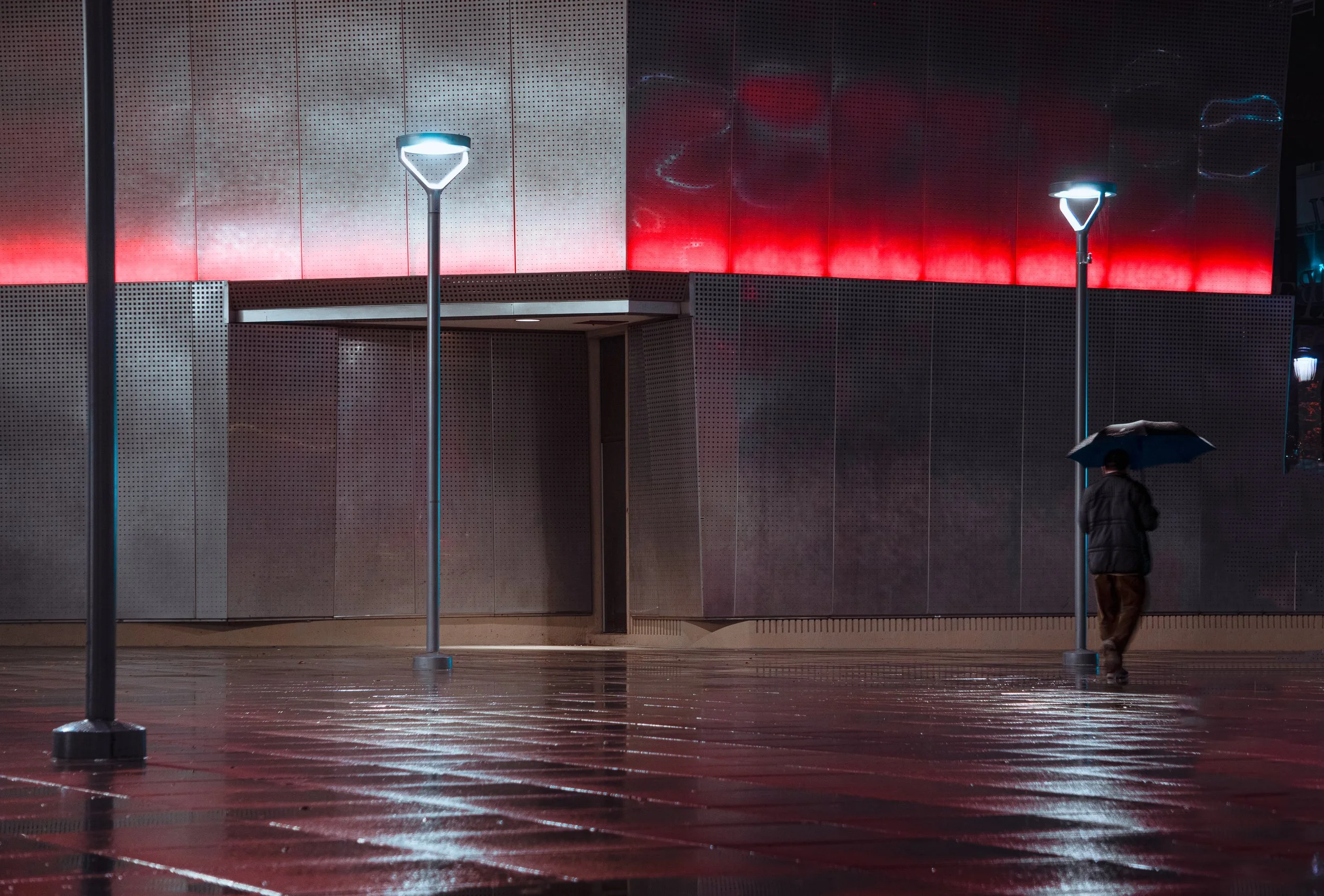 A person with a dark coat and brown pants holding an umbrella walking on wet ground near modern street lamps at night, in front of a building with metallic panels and red and white lighting.