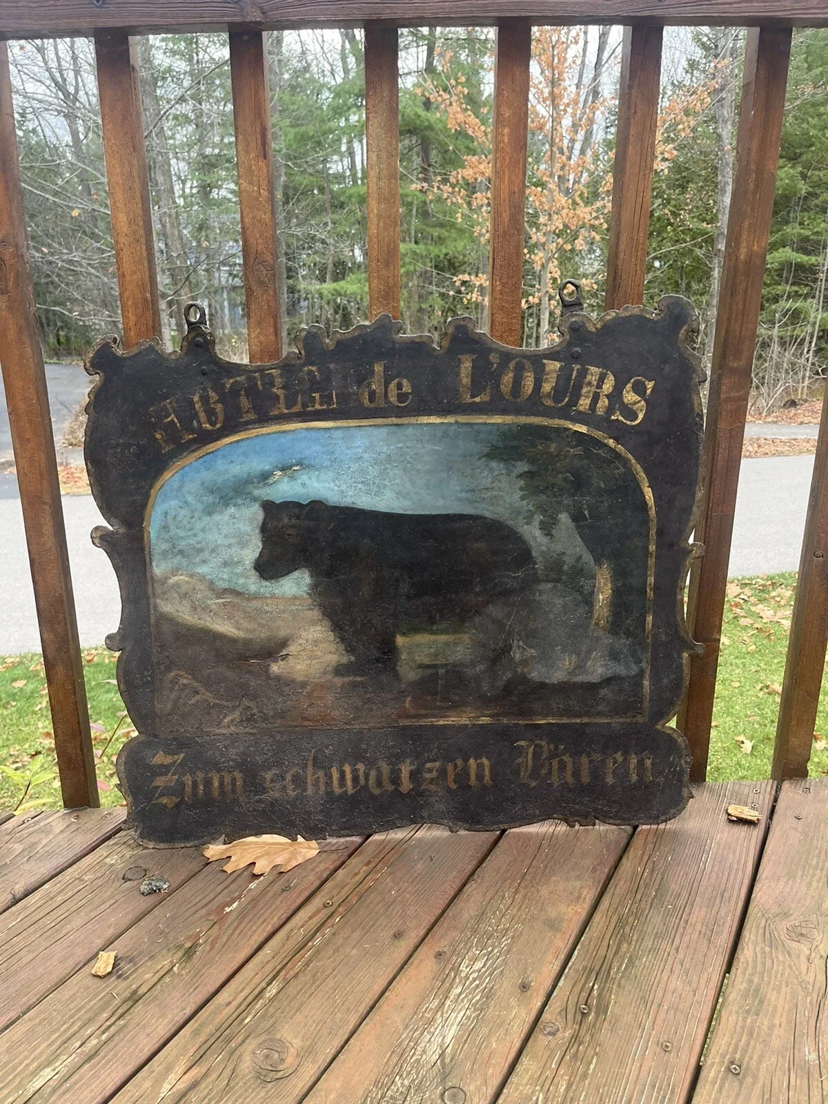 A worn metal sign with a bear illustration in a mountain landscape, displaying French and German text, mounted on a wooden deck railing outdoors.