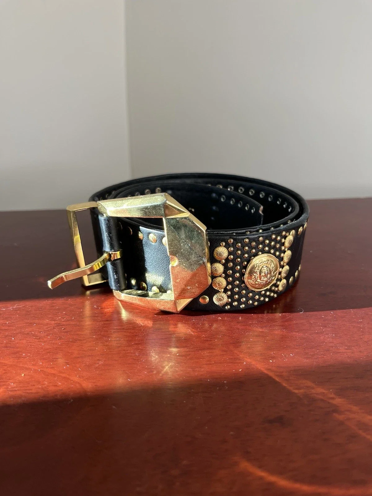 Black leather belt with gold decorative studs and a gold medallion, lying on a wooden surface with a plain wall background.