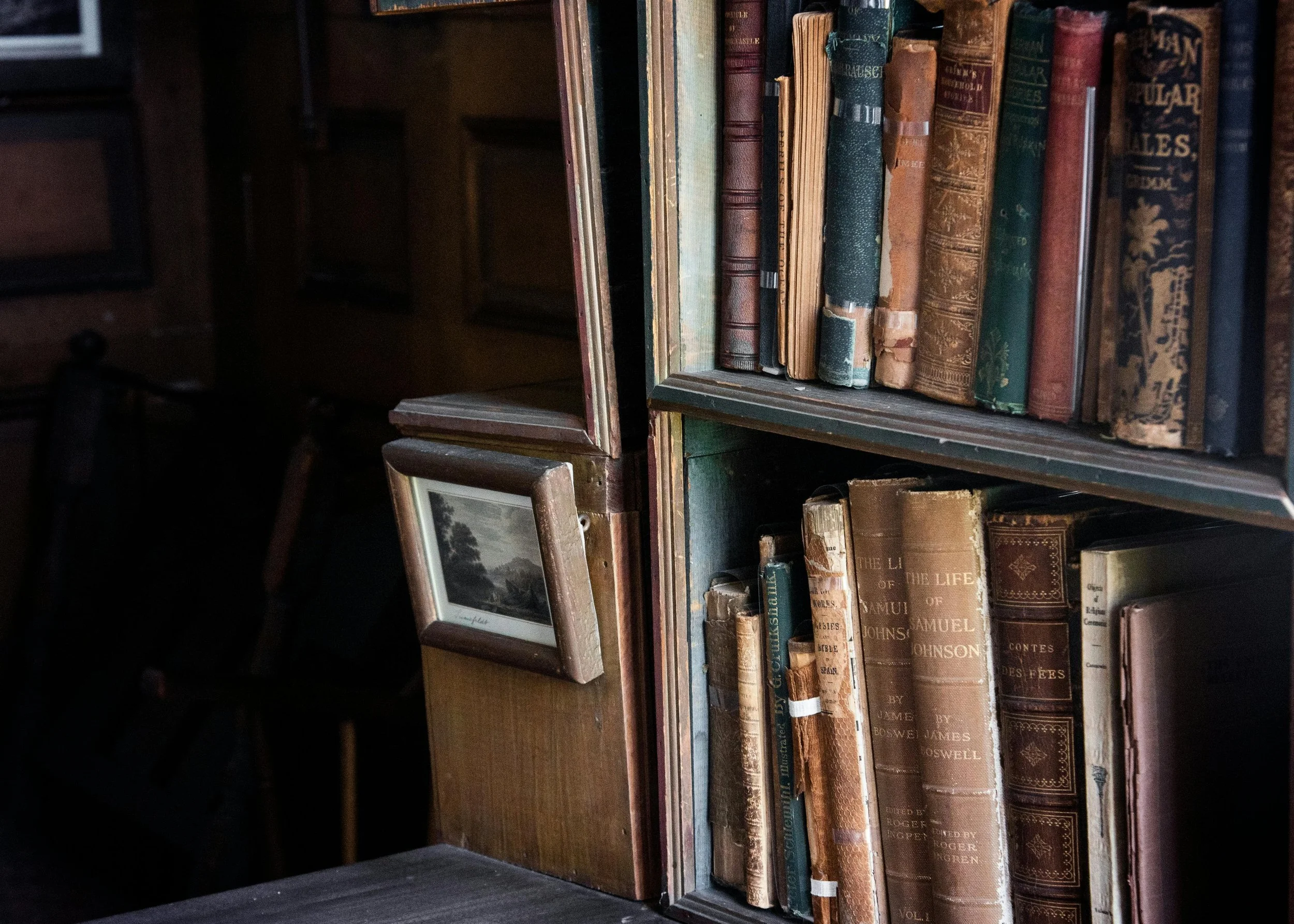 Close-up of a wooden bookshelf filled with old, worn books, with a small framed landscape picture leaning against it, in a dimly lit room.