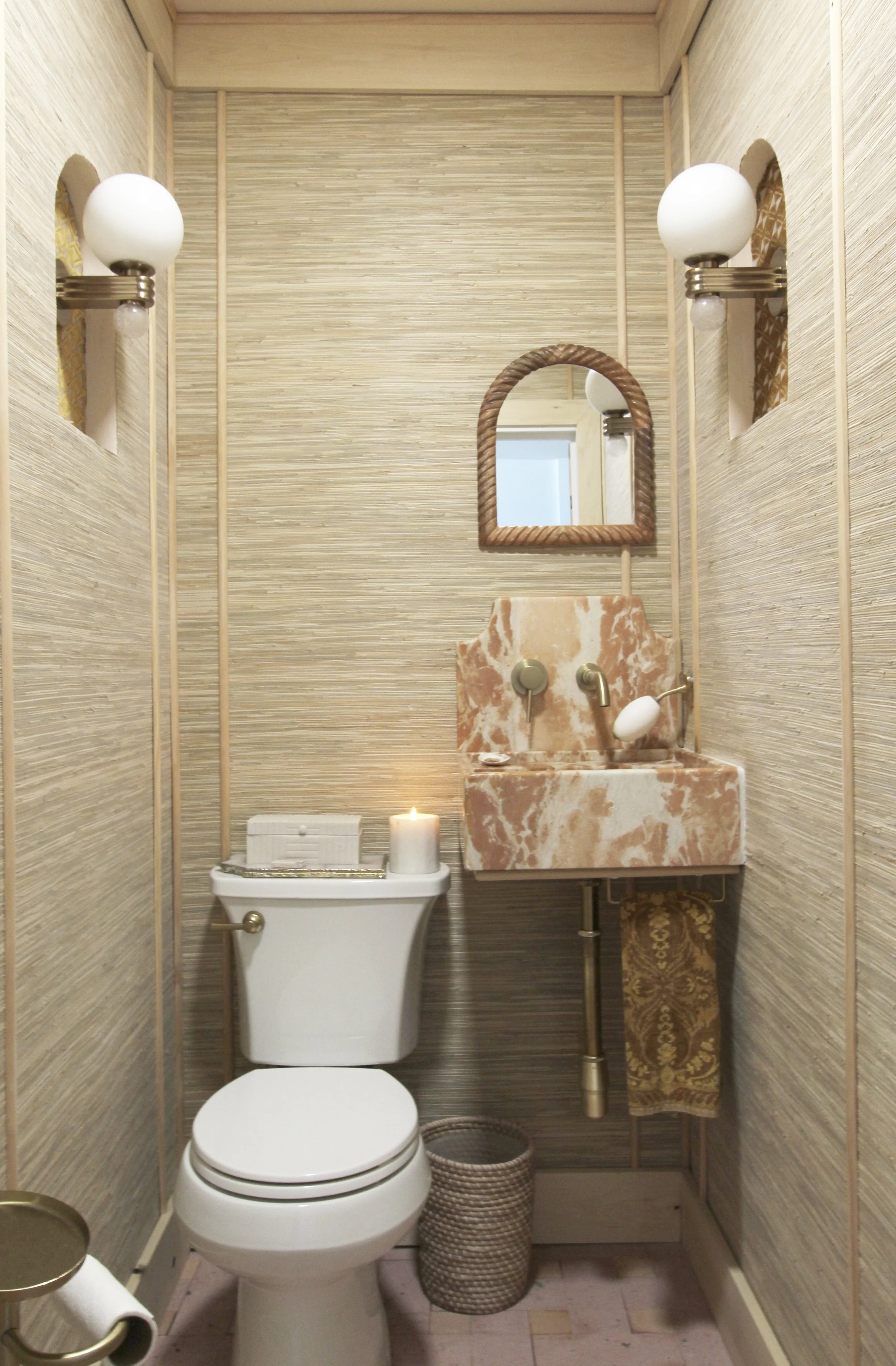 A small bathroom with beige textured walls, a white toilet with a lit candle on top, a pink and white marble sink with brass fixtures, a small arched mirror above the sink, two wall sconces with white globes on either side, a small woven basket on th
