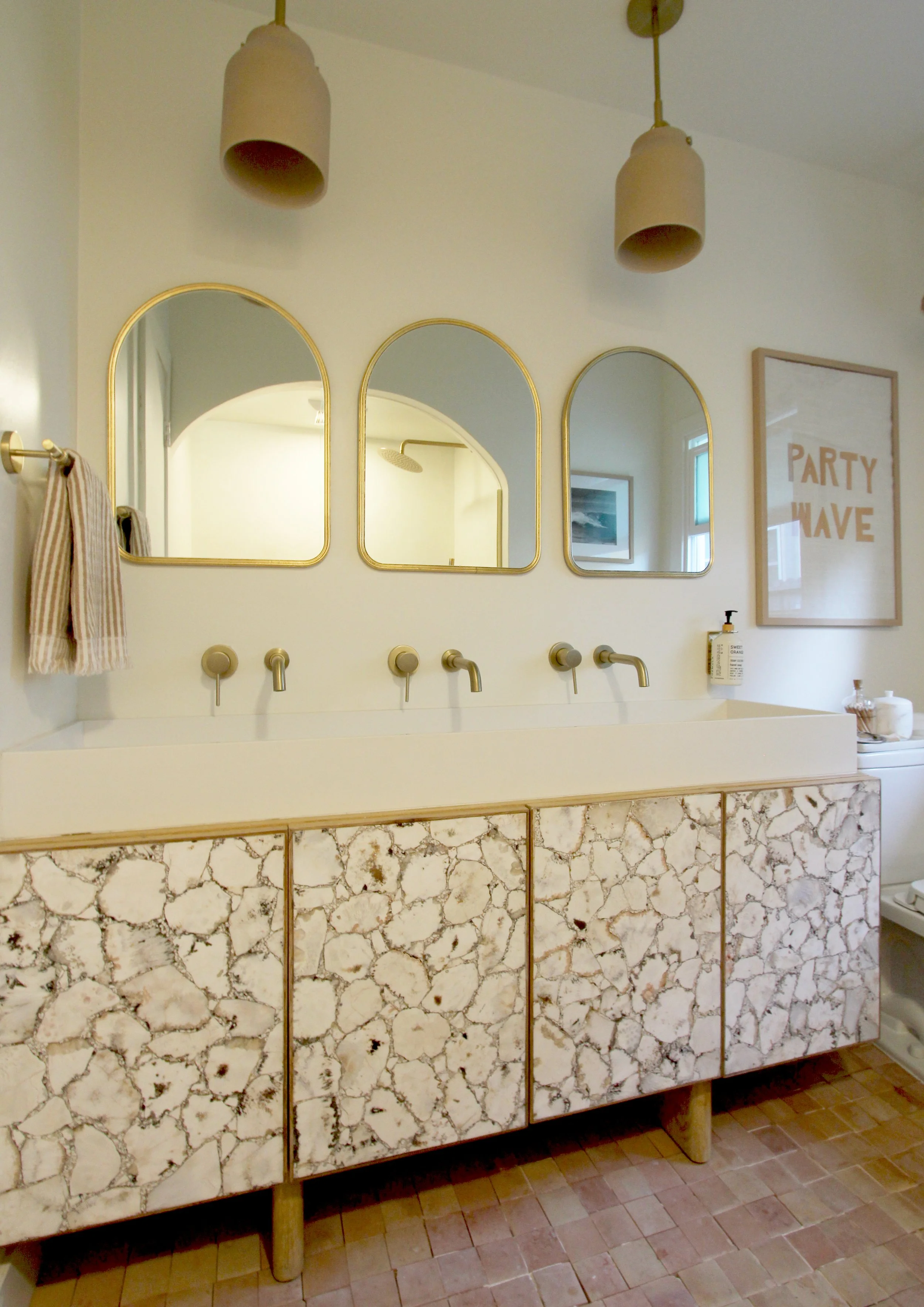 A bathroom vanity with a white countertop and a marble-patterned cabinet with gold edges. Four beige faucets are on the countertop, with round mirrors above them. Two beige pendant lights hang from the ceiling, and a framed sign that reads "PARTY WAV