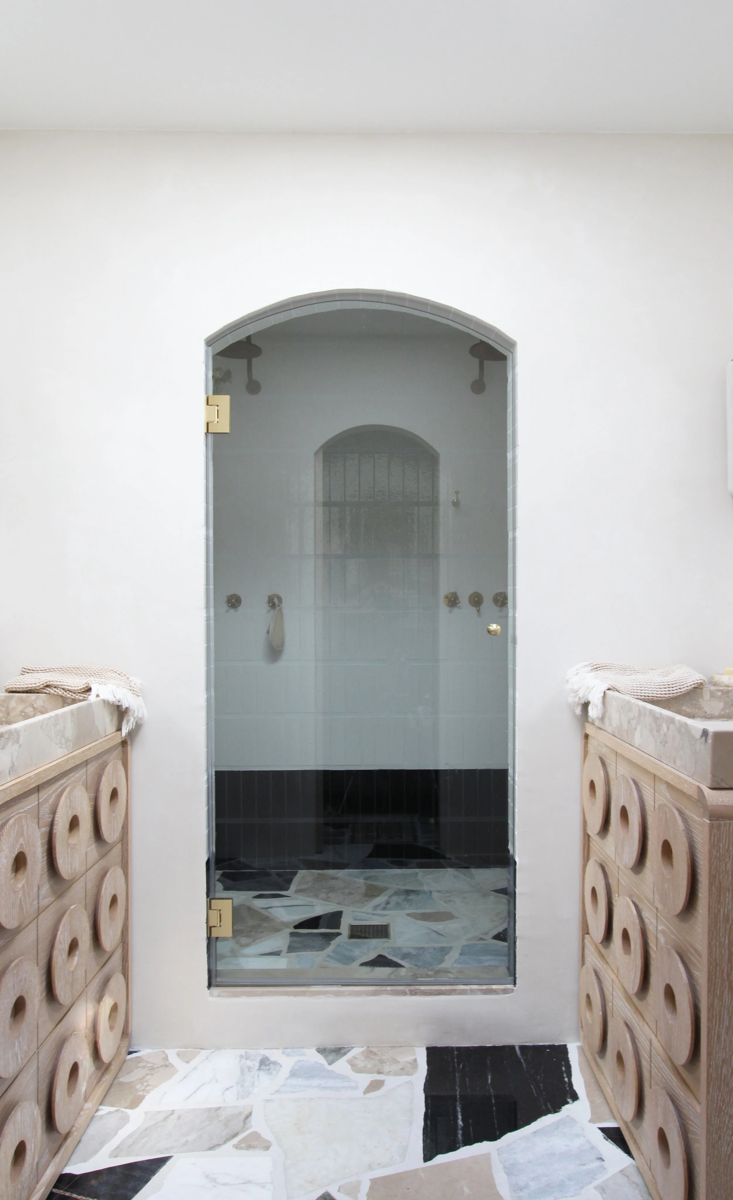 Bathroom with a walk-in shower at the center, framed with glass door, flanked by wooden cabinets with rounded cutouts, and a geometric tile floor.