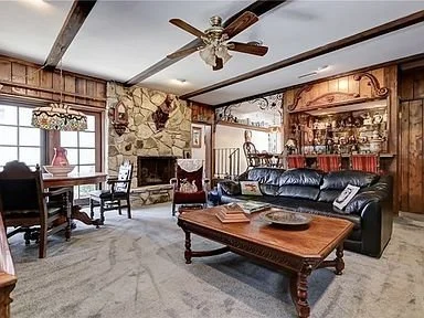 Living room with wooden paneling, stone fireplace, ceiling fan, leather sofa, wooden coffee table, dining table, and large windows.