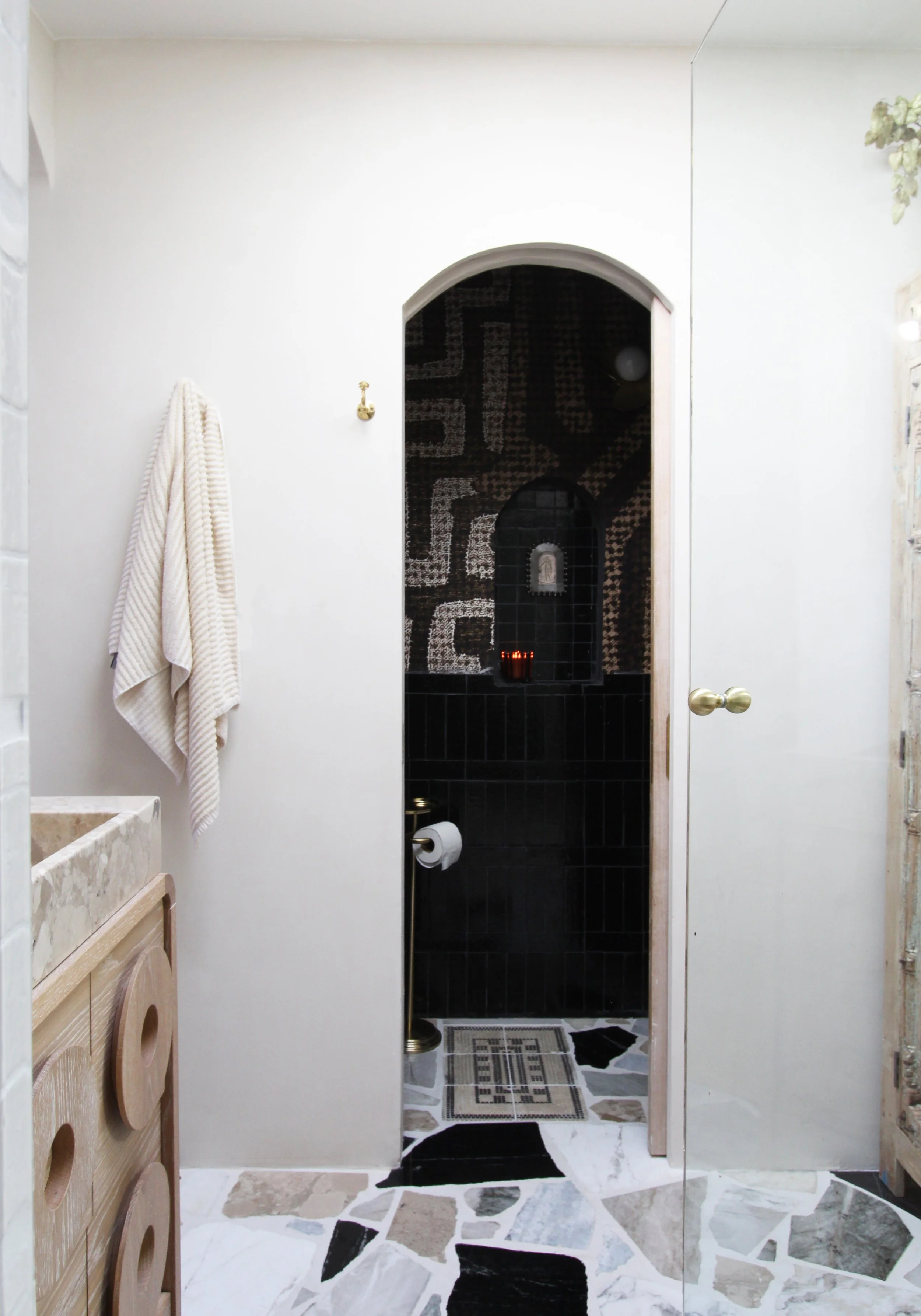 Bathroom with white walls, a beige towel hanging on a hook, a marble sink on a wooden cabinet, and a doorway leading to a black-tiled shower area with dark mosaic wall tiles and a decorative pattern, with a patterned floor outside the shower.