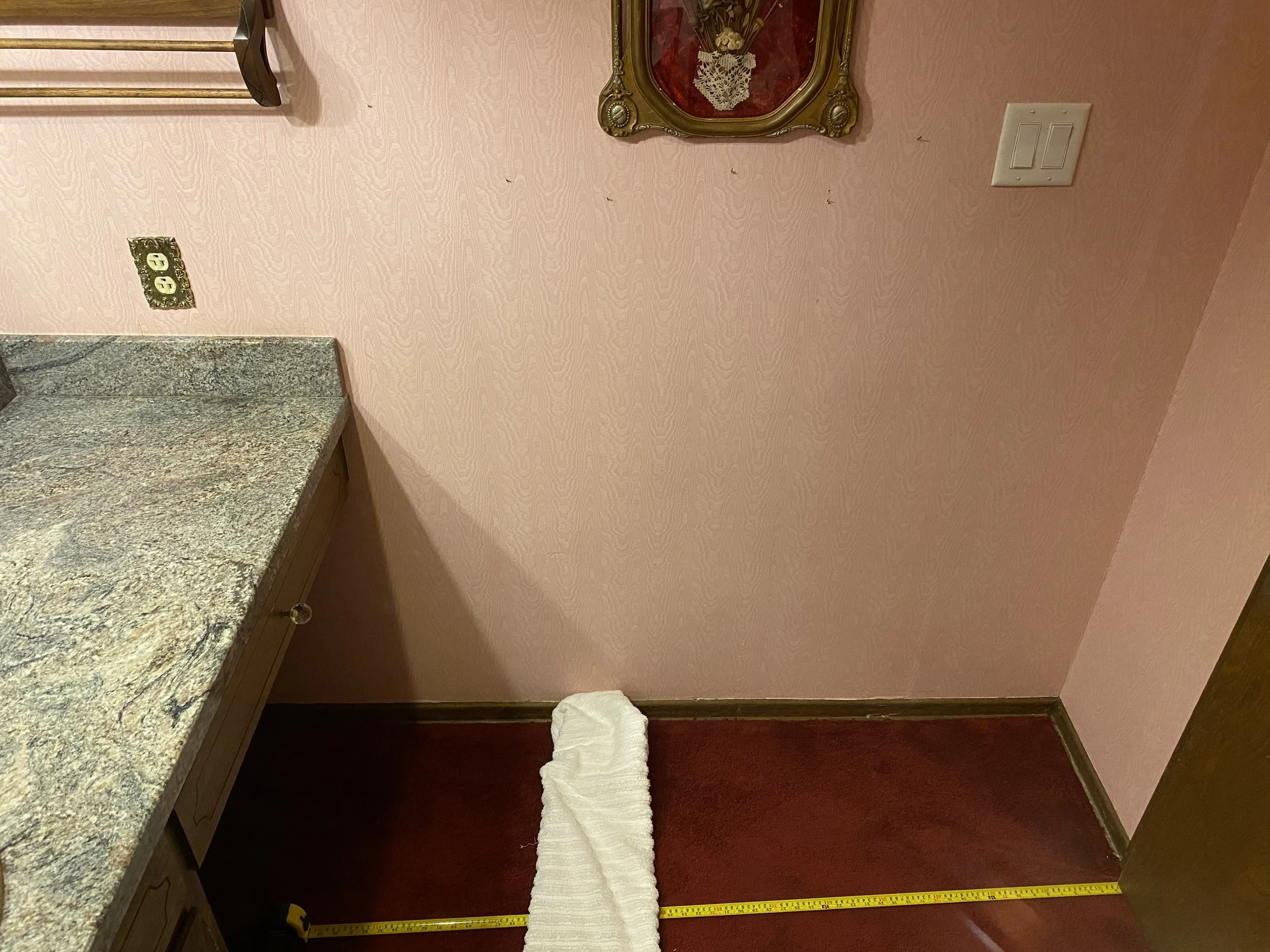Corner of a room with a pink wall, a granite countertop, and a measuring tape on the floor, with a rolled paper towel on the floor, a picture frame on the wall, and a light switch.