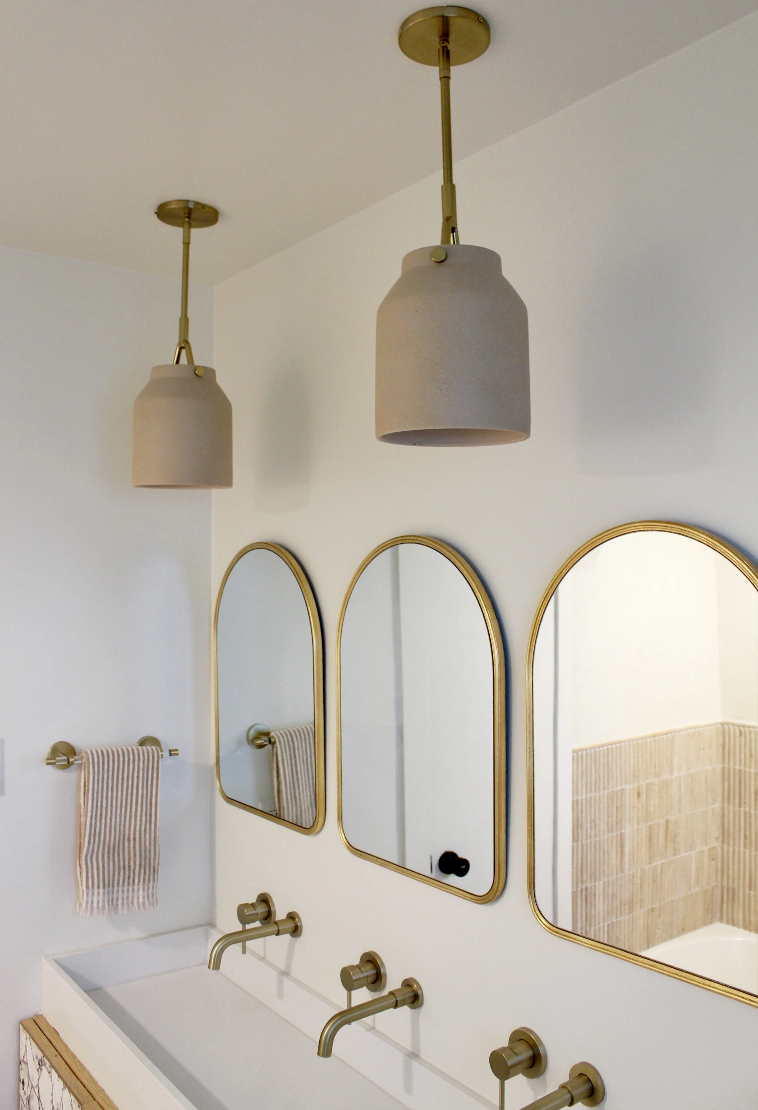 Modern bathroom with three arched mirrors with gold frames, two pendant lights, a gold faucet, and a striped towel on a towel rack.