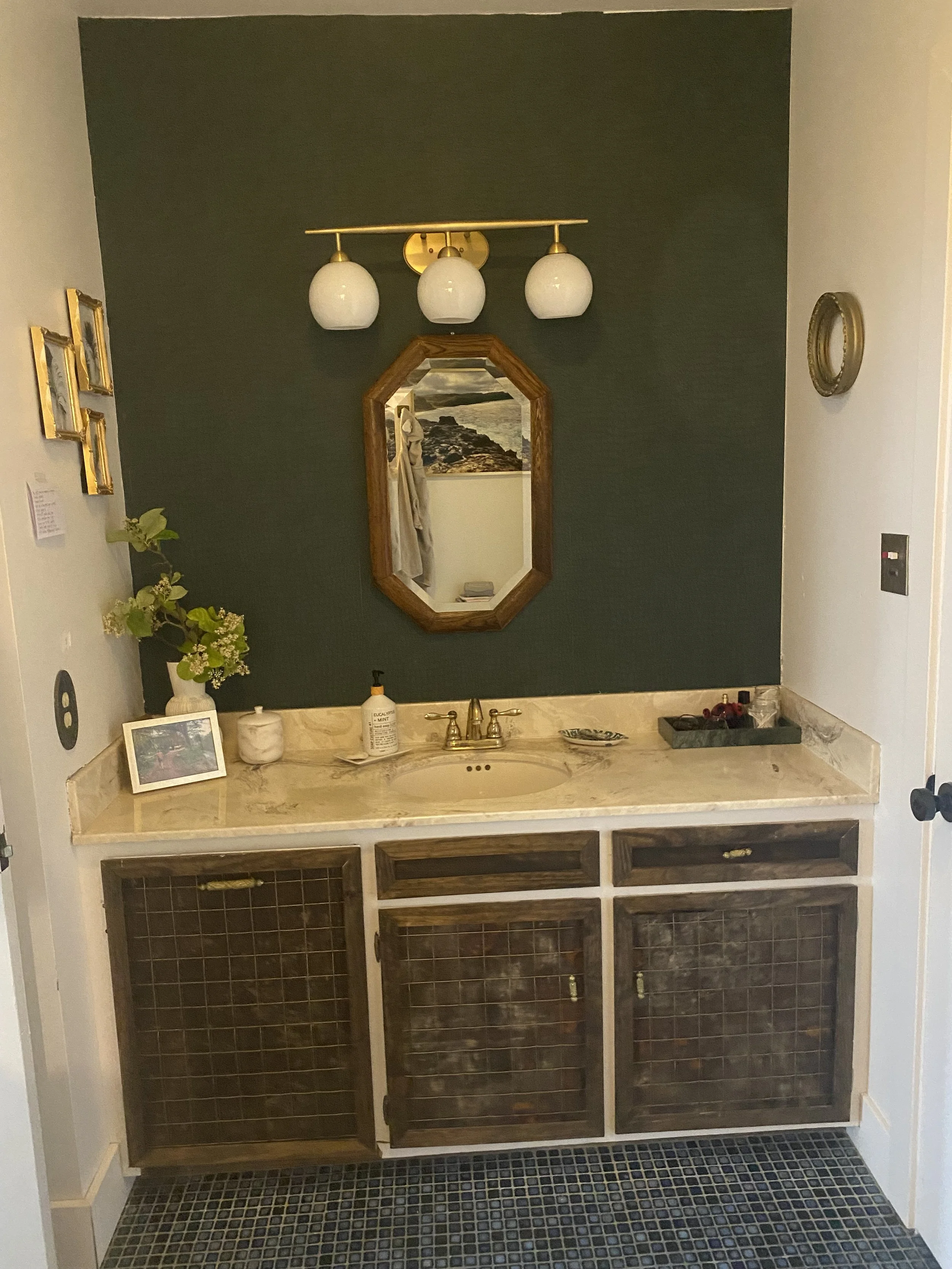 Bathroom vanity with a marble countertop, gold faucet, decorative plant, framed photo, bottle of hand sanitizer, and tray with red and purple items. A vertical mirror, wall-mounted light fixture with three globe-shaped bulbs, and wall decorations are