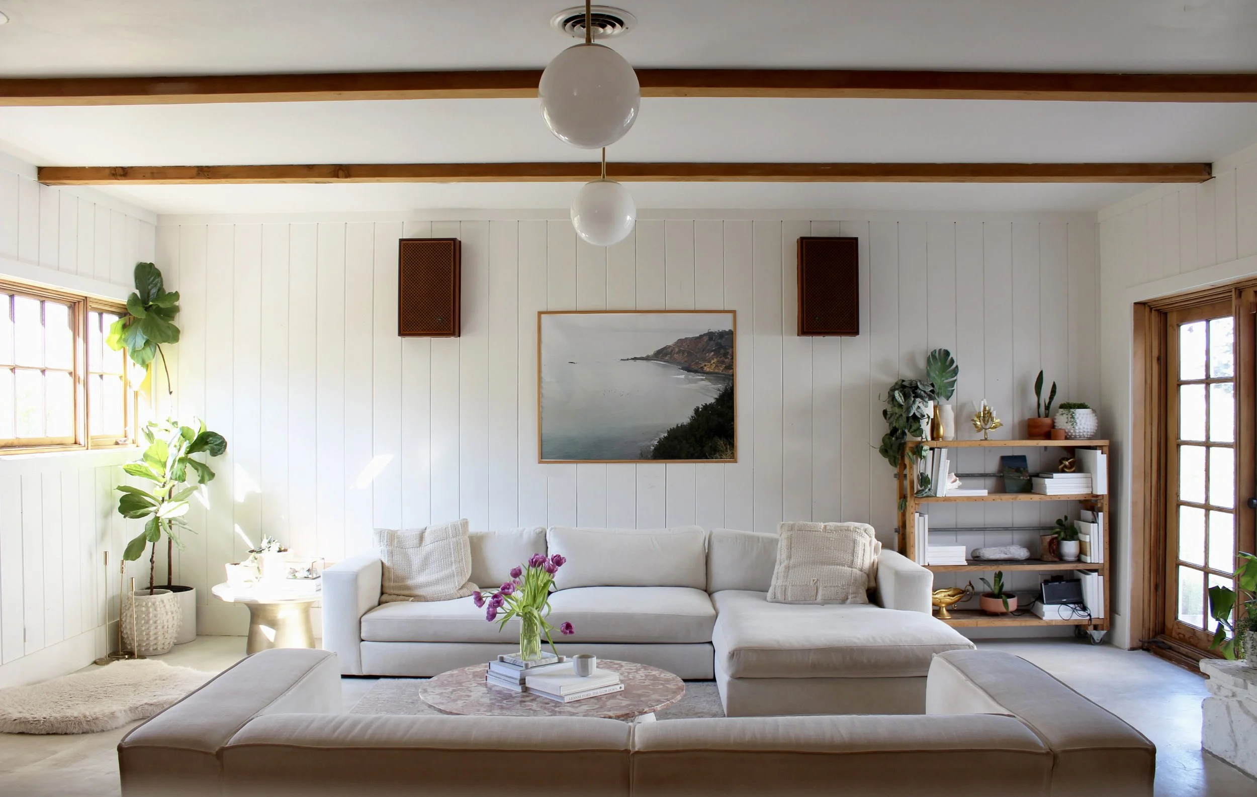 Bright living room with beige sectional sofa, coffee table with pink flowers, white wall paneling, window with wooden trim, bookshelf with plants and decor, large artwork of a coastal landscape, and ceiling lights.