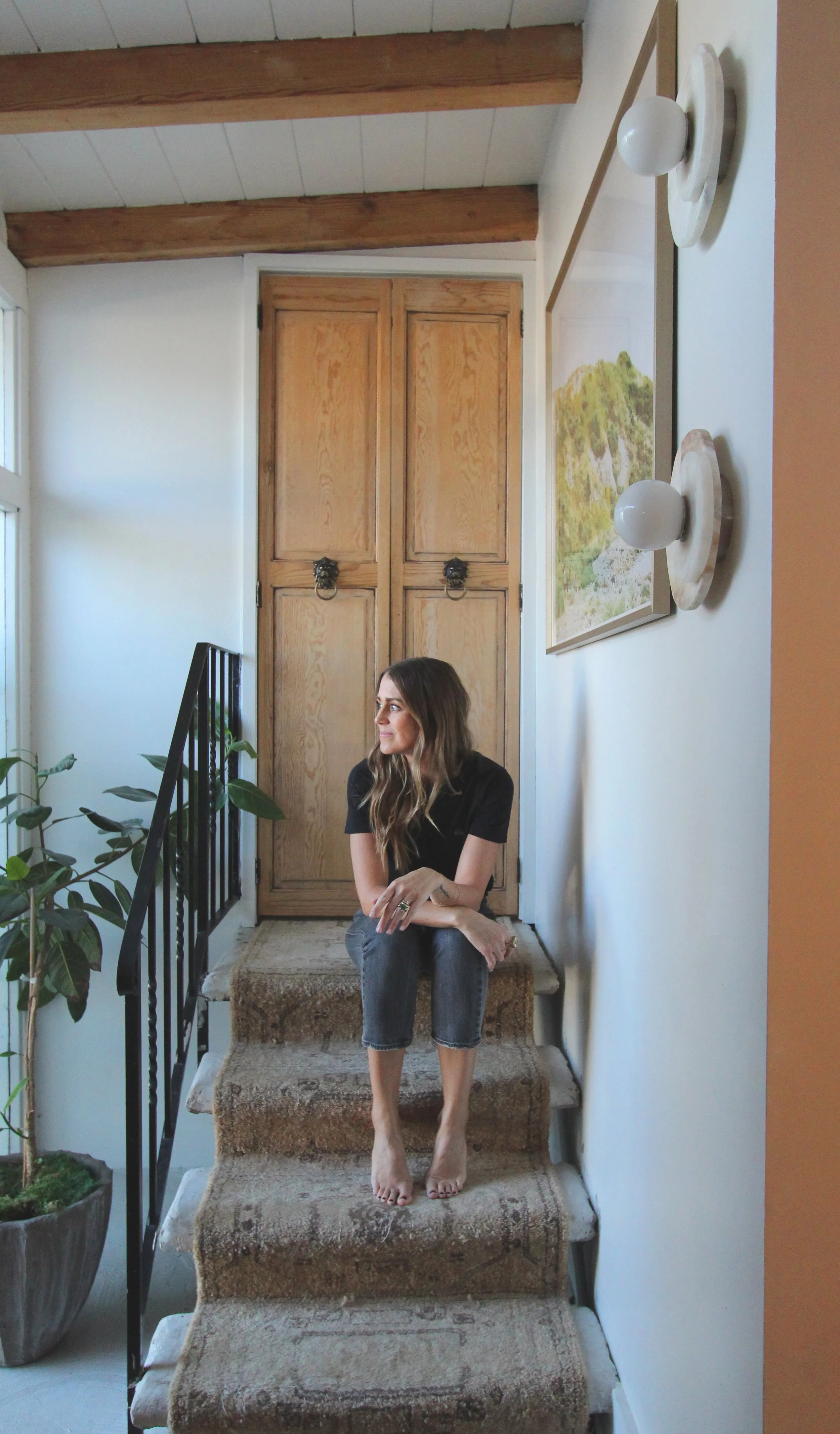A woman in a black shirt and rolled-up jeans sits on carpeted stairs in a sunny, rustic interior. There are wooden cabinets behind her, a large potted plant on her left, and a wall-mounted painting above. The ceiling has exposed wooden beams, and a round modern light fixture is visible.