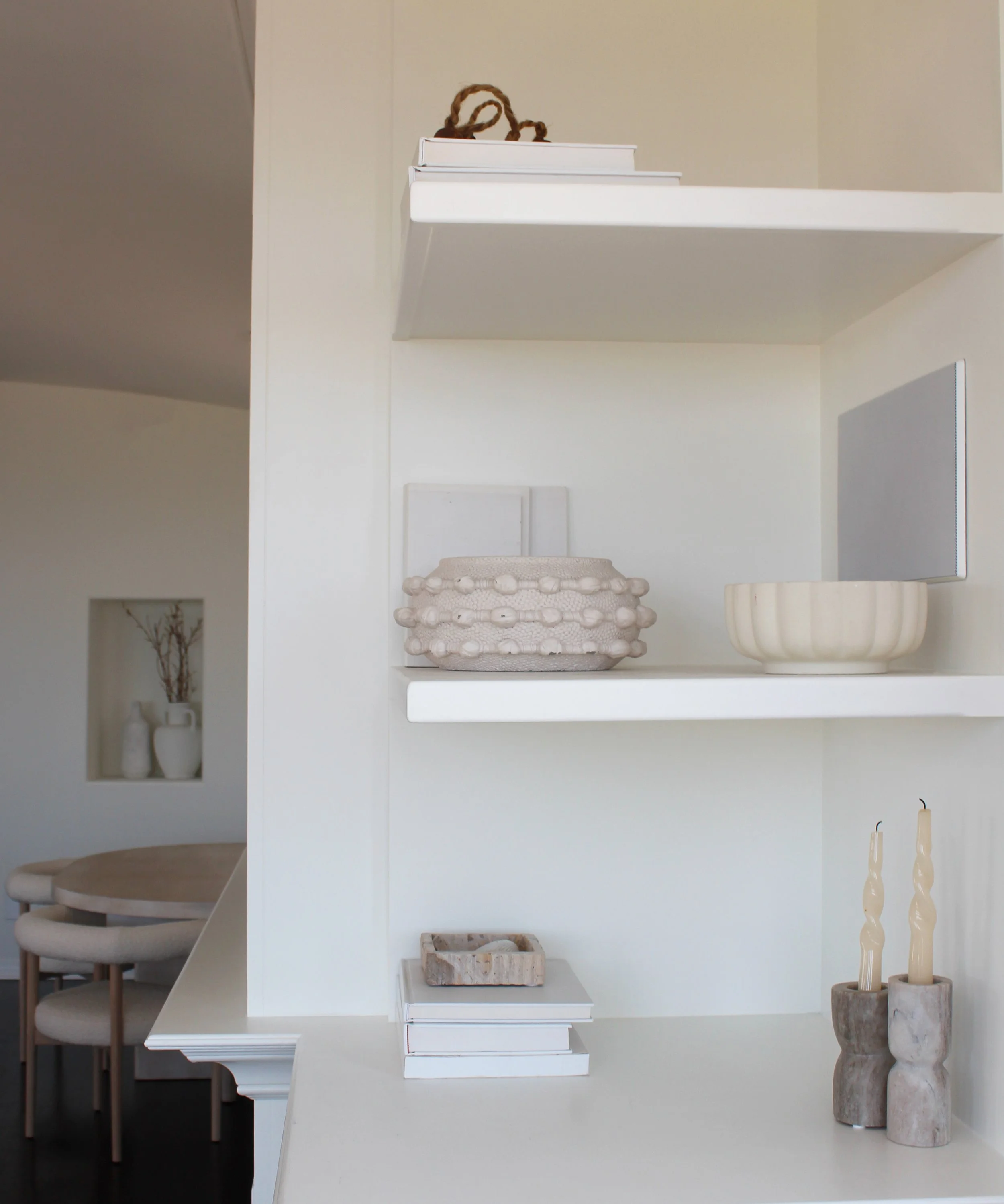 White wall with floating shelves holding decorative objects, including vases, bowls, and small books, with a dining area with beige chairs in the background.