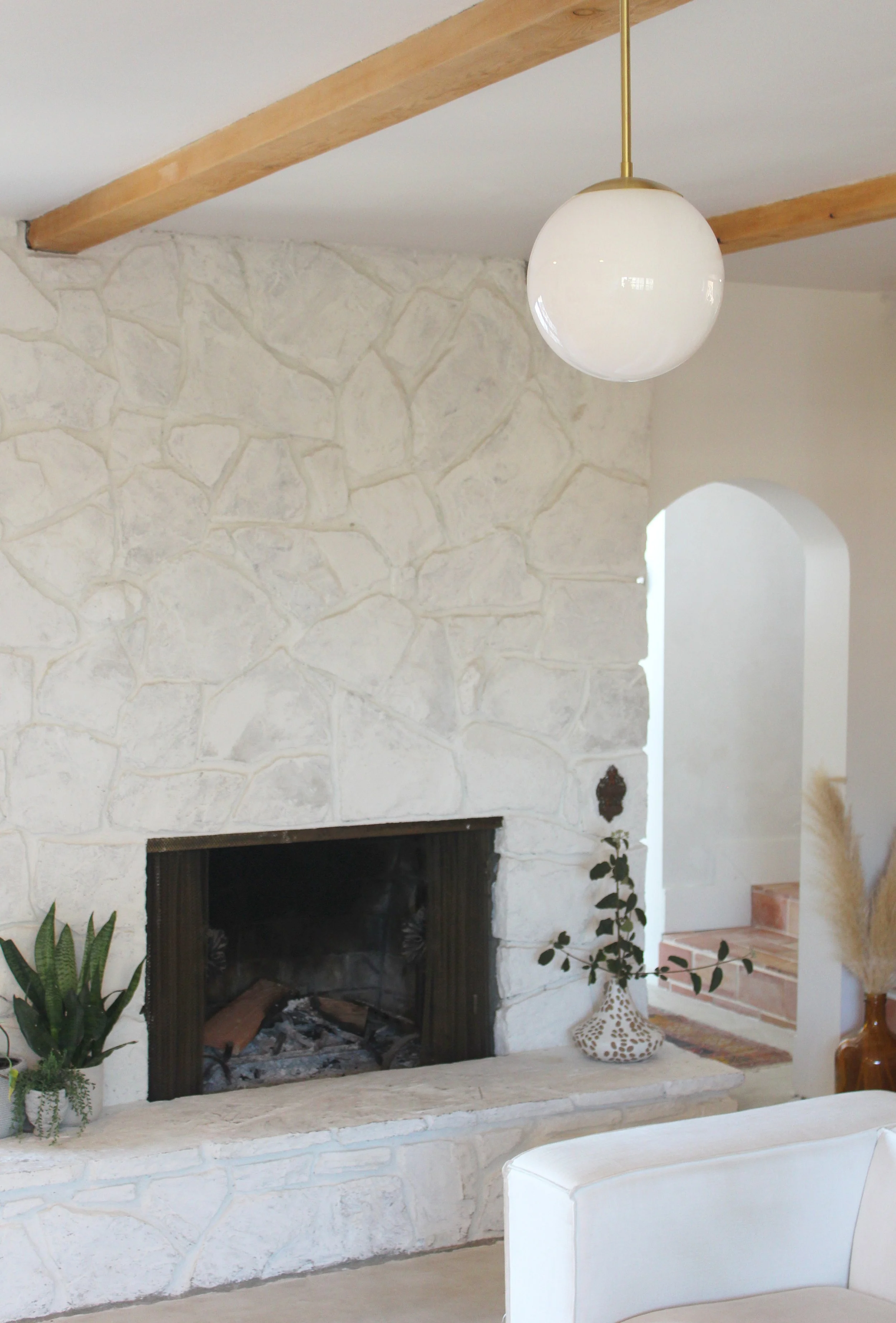 Living room with a white stone fireplace, a round white pendant light, a green plant in a decorative vase, and a staircase with pinkish tiles in the background.