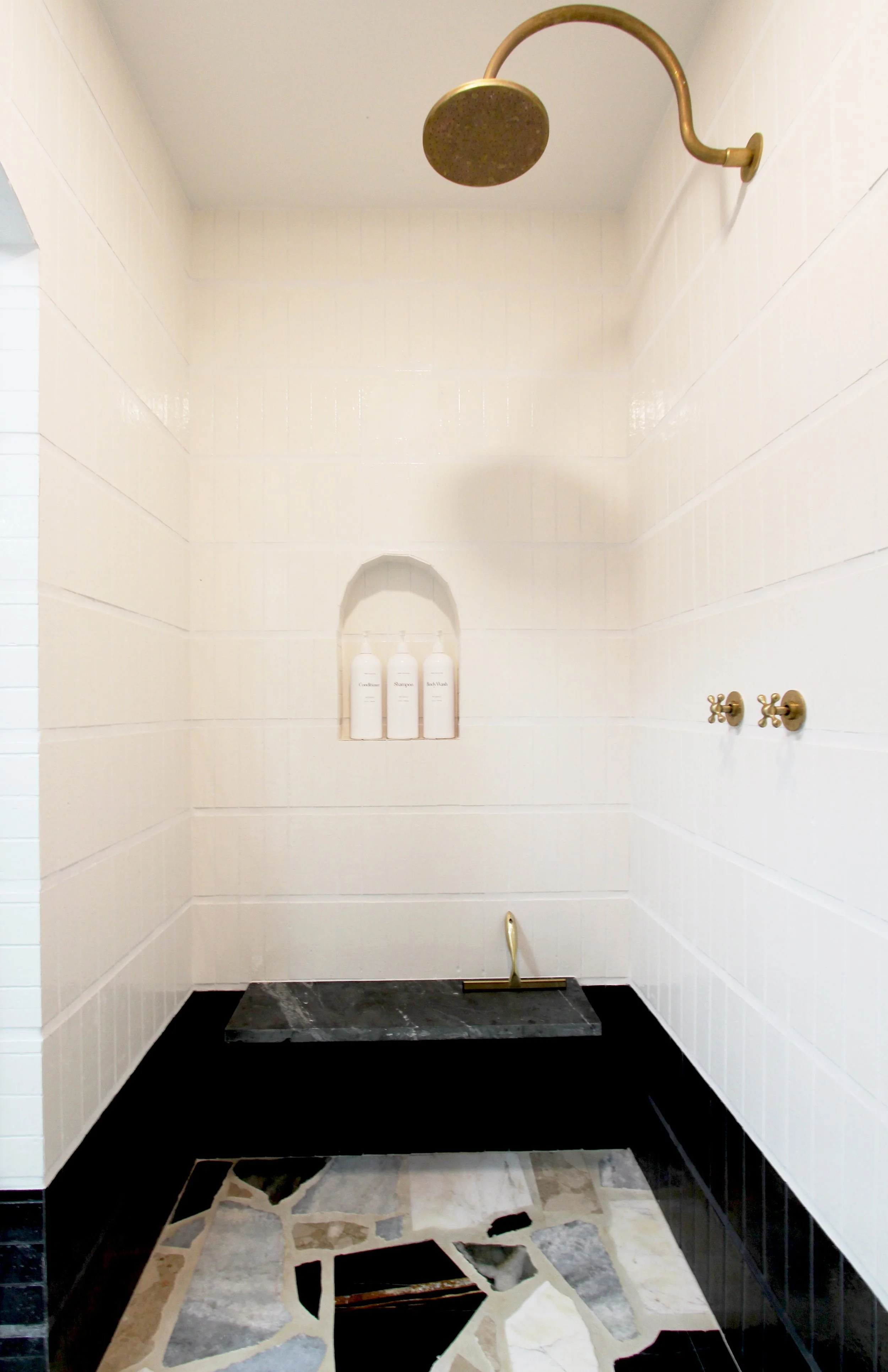 Shower area with a gold showerhead and handles, beige tiled walls, a small niche with bottles, black marble shower base, and an irregular patterned stone floor.