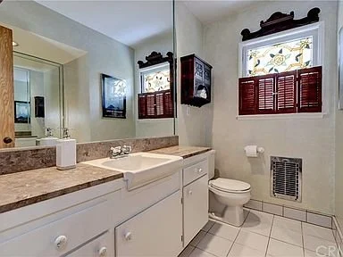 Bathroom with white vanity, marble countertop, sink, mirror, small window with decorative wood shutters, toilet, wall-mounted heater, and tiled floor.