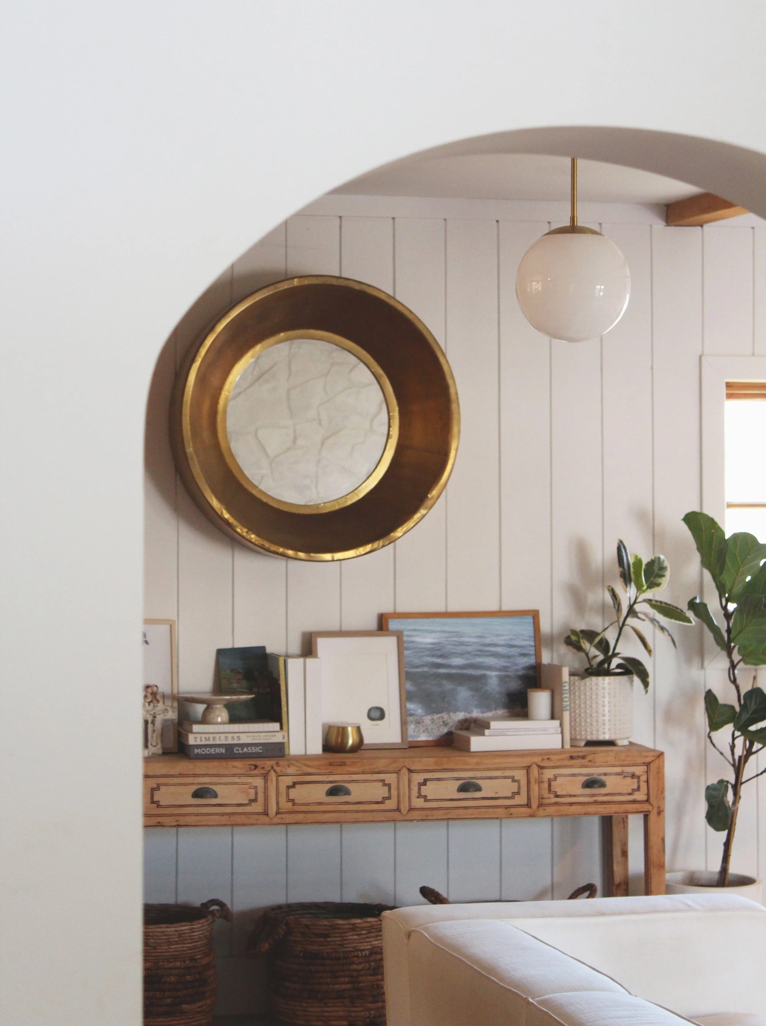 Living room with a wooden console table decorated with framed pictures, books, a candle, and a small gold container. There's a large round mirror with a gold frame on the wall and a hanging globe light fixture. A potted plant with large green leaves is on the right side. Baskets are underneath the console table, and there's a white upholstered sofa in the foreground.