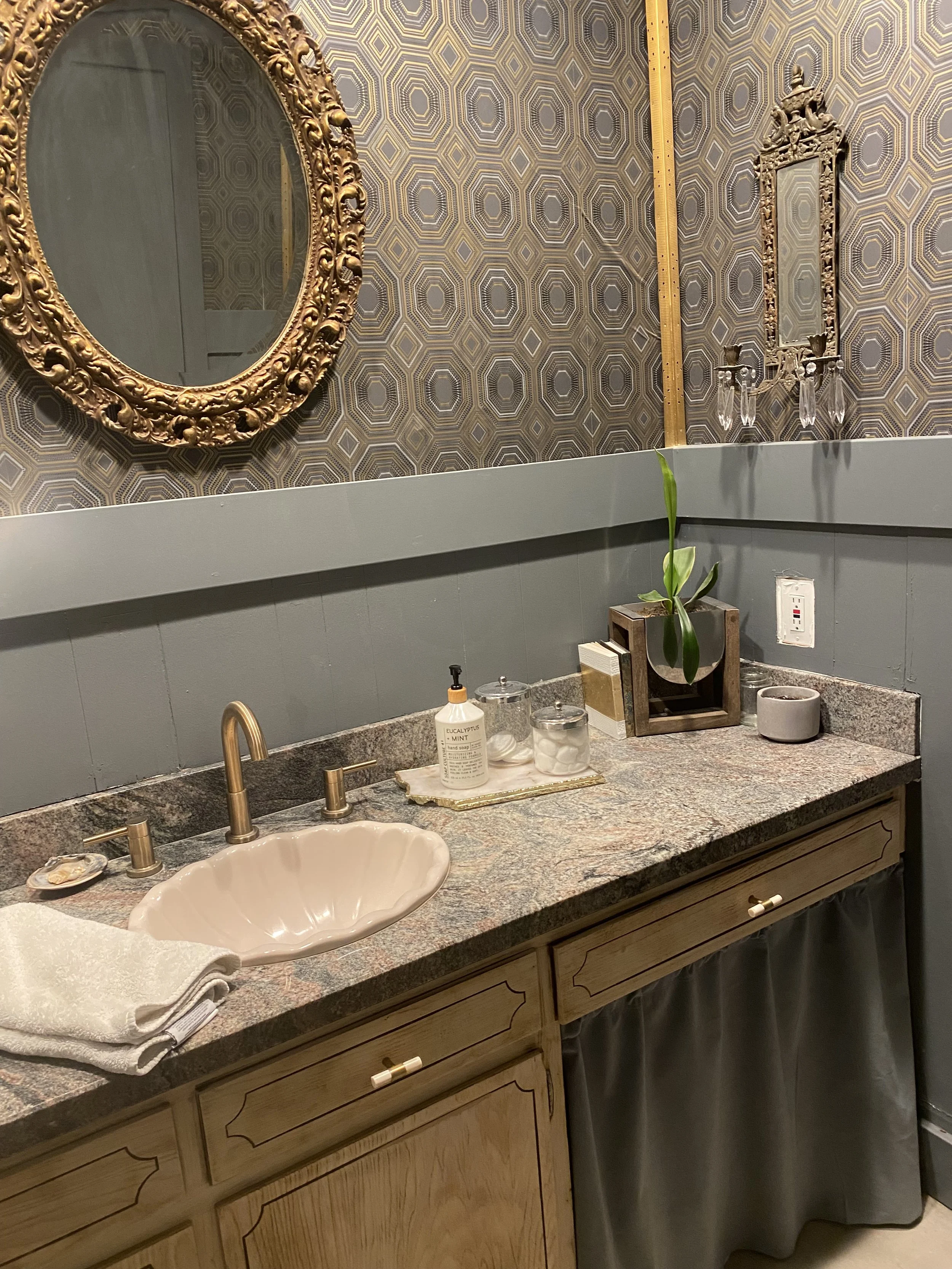 Bathroom vanity with a pink sink, golden faucet, and a granite countertop. On the countertop are soap, lotion, jars, a plant in a wooden holder, and a small bowl. A mirror with an ornate gold frame hangs on the decorative patterned wall.