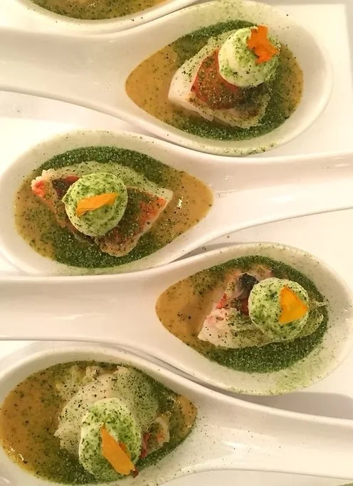 Four white porcelain tasting spoons topped with green foam and small orange garnishes, arranged on a white platter with green sauce or herb oil.
