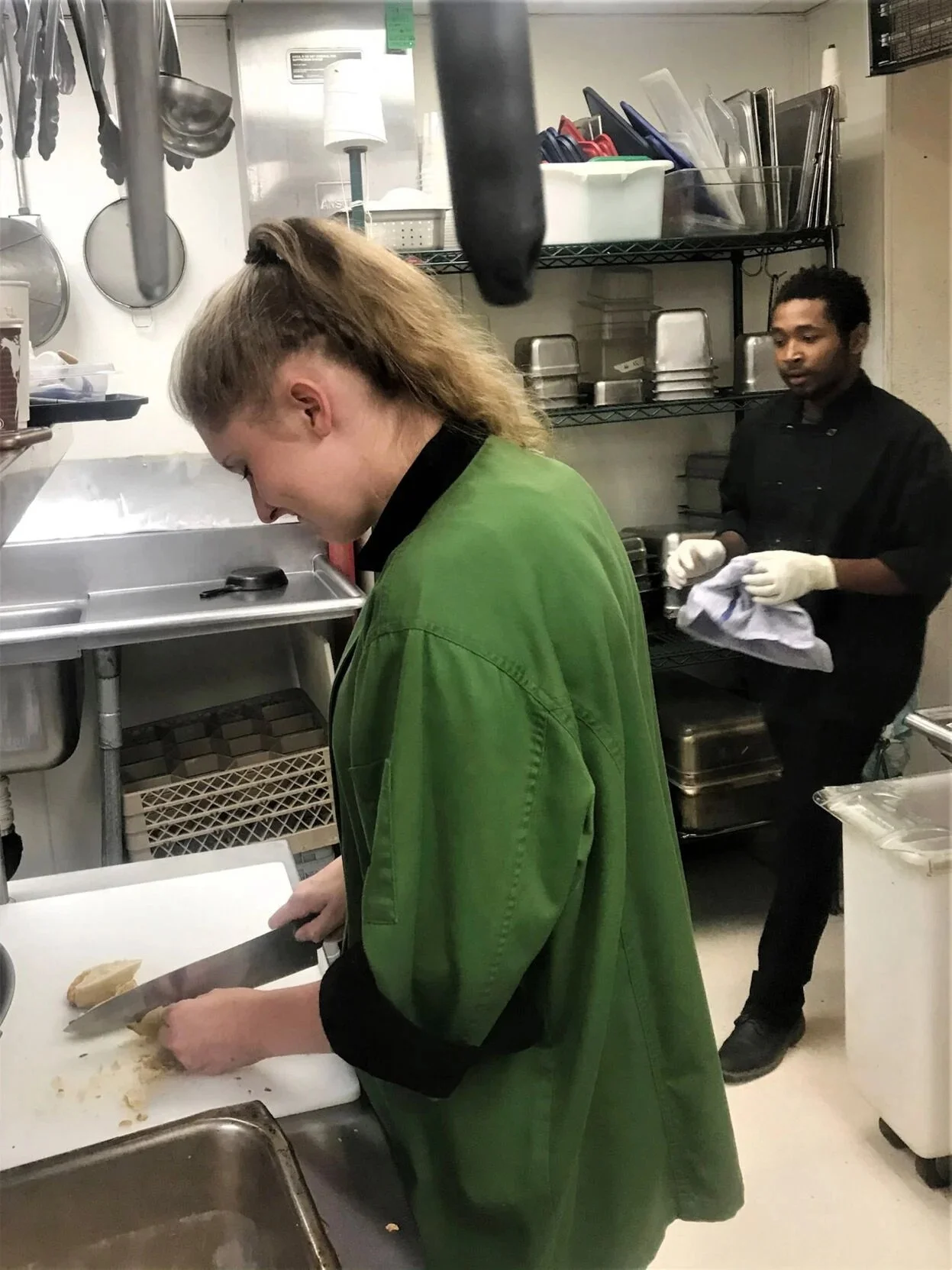 A female chef chopping ginger in a commercial kitchen with a male chef in the background holding a towel.