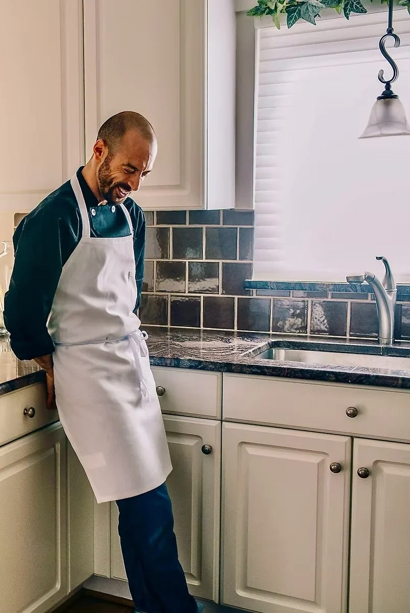 A man wearing a white apron is smiling and looking down in a kitchen with white cabinets, gray tile backsplash, and a window with blinds, near a sink.